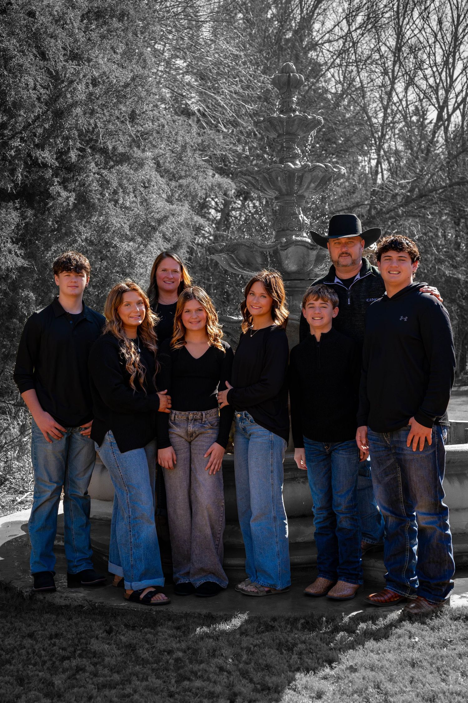 Family of eight in matching black tops and jeans poses together in front of a stone fountain, with one man wearing a cowboy hat.