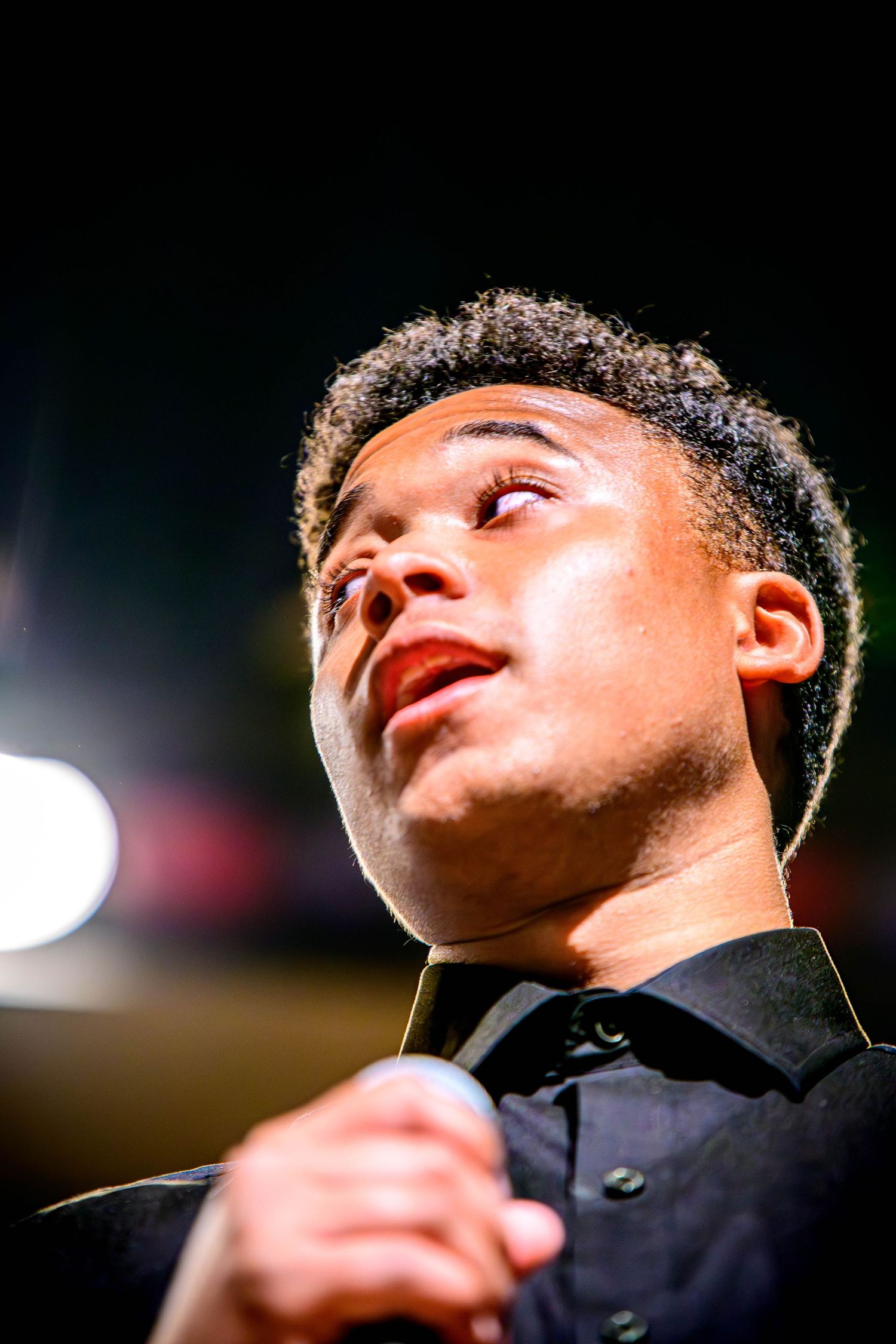 Young man in a black button-up shirt gazes upward while holding a microphone during a live performance under dramatic stage lighting.