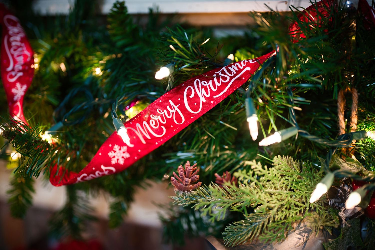 Red Merry Christmas ribbon decorates a green artificial Christmas tree with glowing white lights.