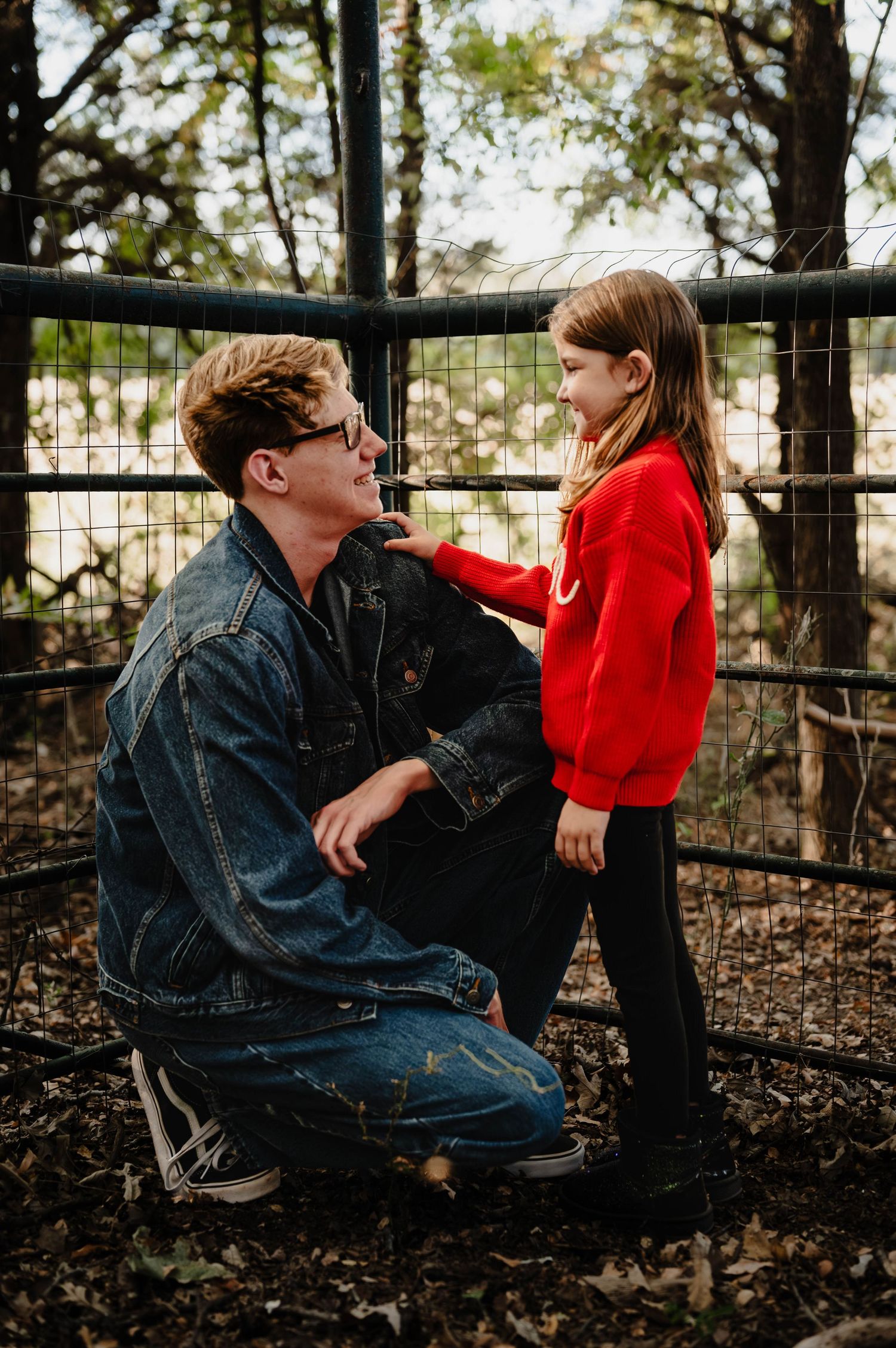 A child in a bright red sweater stands next to an adult in denim against a rustic fence background. Azle family photo session.