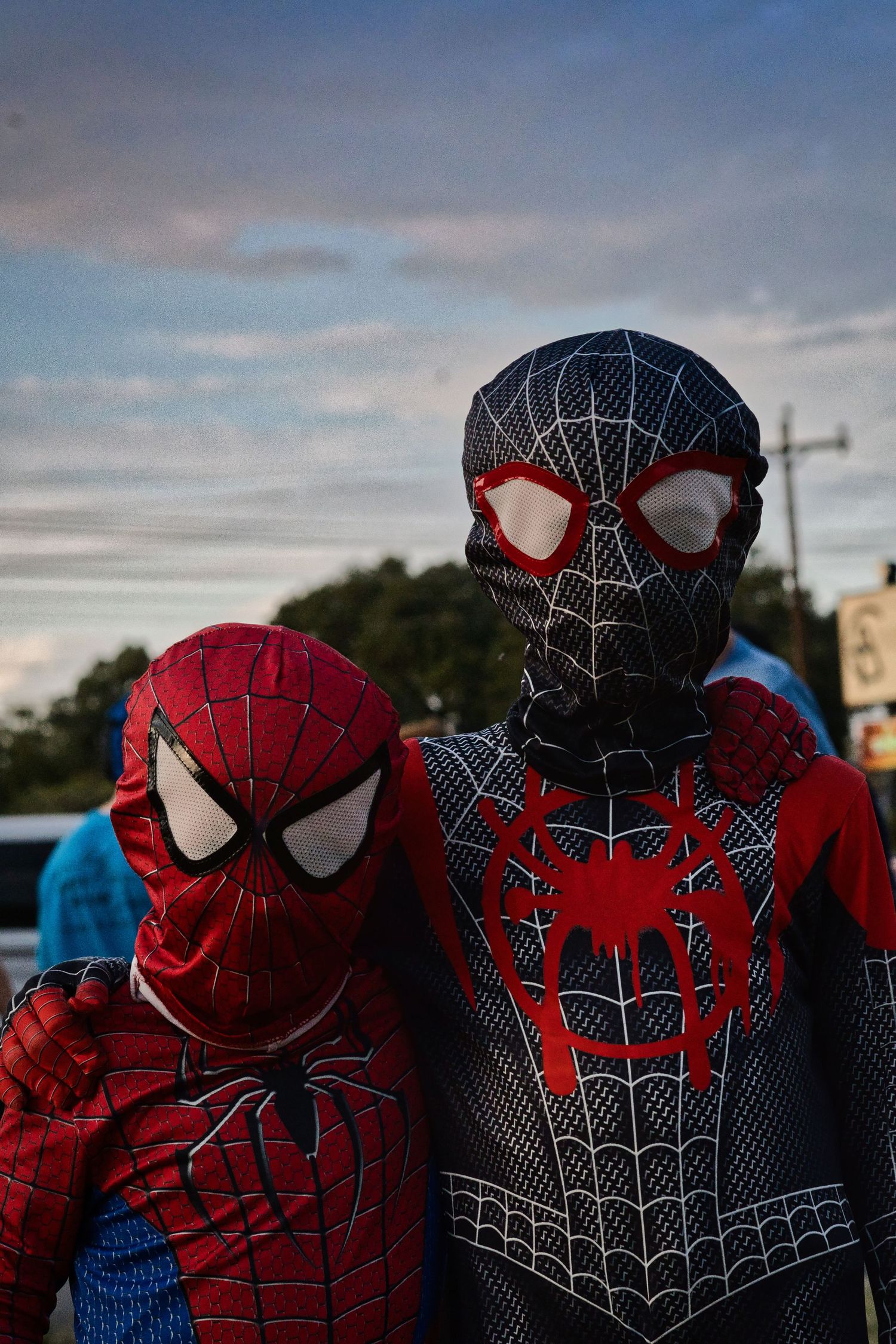 Two people in Spider-Man costumes, one in classic red and blue and one in a black and red suit, stand together at dusk.