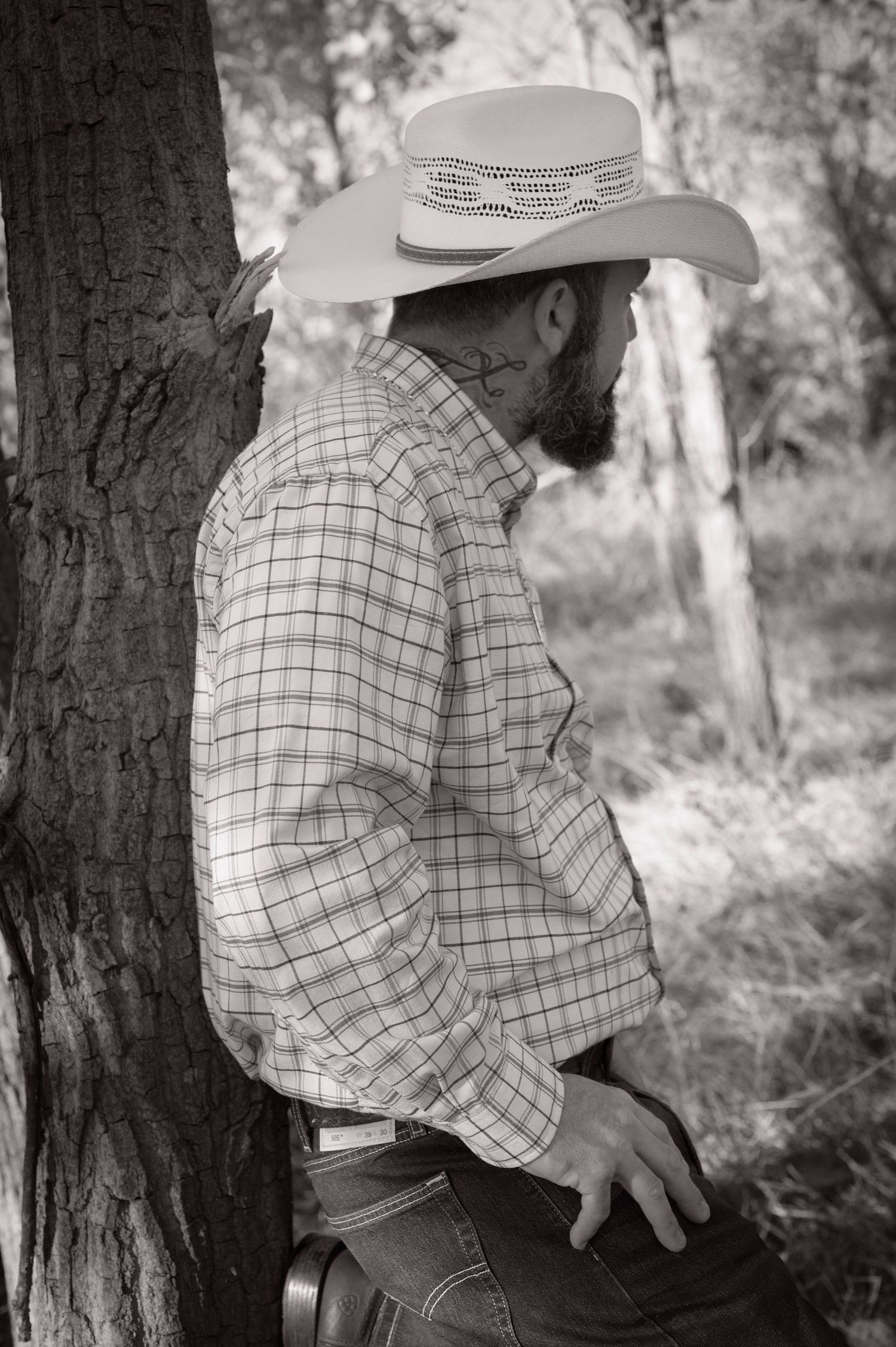 A person in a plaid shirt and cowboy hat sits on a bench in a black and white forest setting. Azle family photo session.