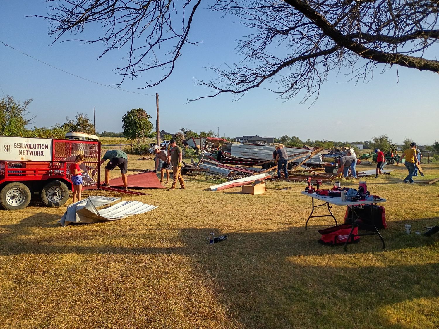 Outdoor aviation event with people assembling glider aircraft components on grass near a red trailer on a sunny day.