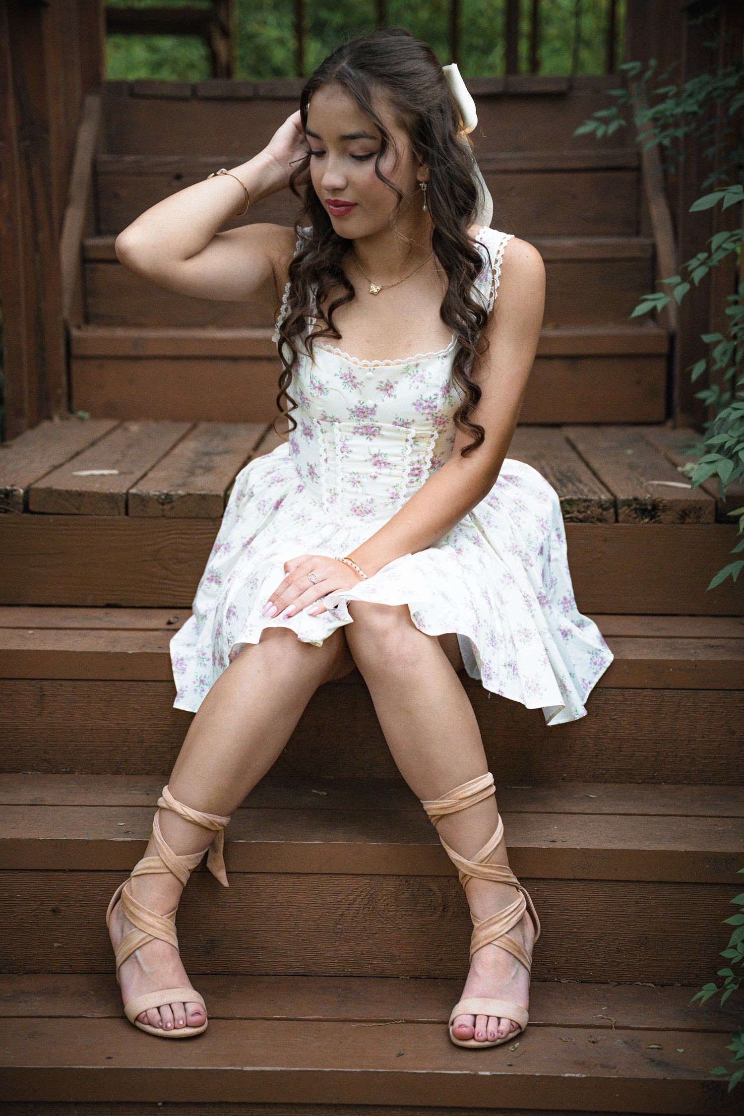 Young woman in floral dress and strappy heels sitting gracefully on wooden steps outdoors.