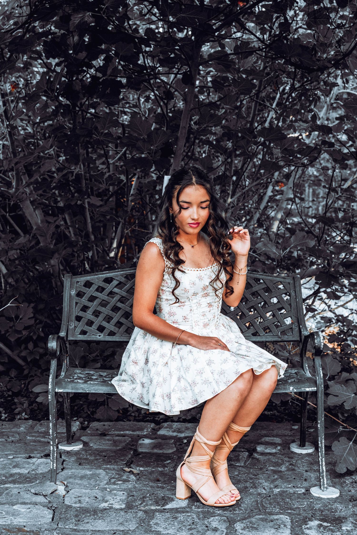 Young woman in floral dress and strappy heels sitting gracefully on an ornate metal bench surrounded by dark foliage.