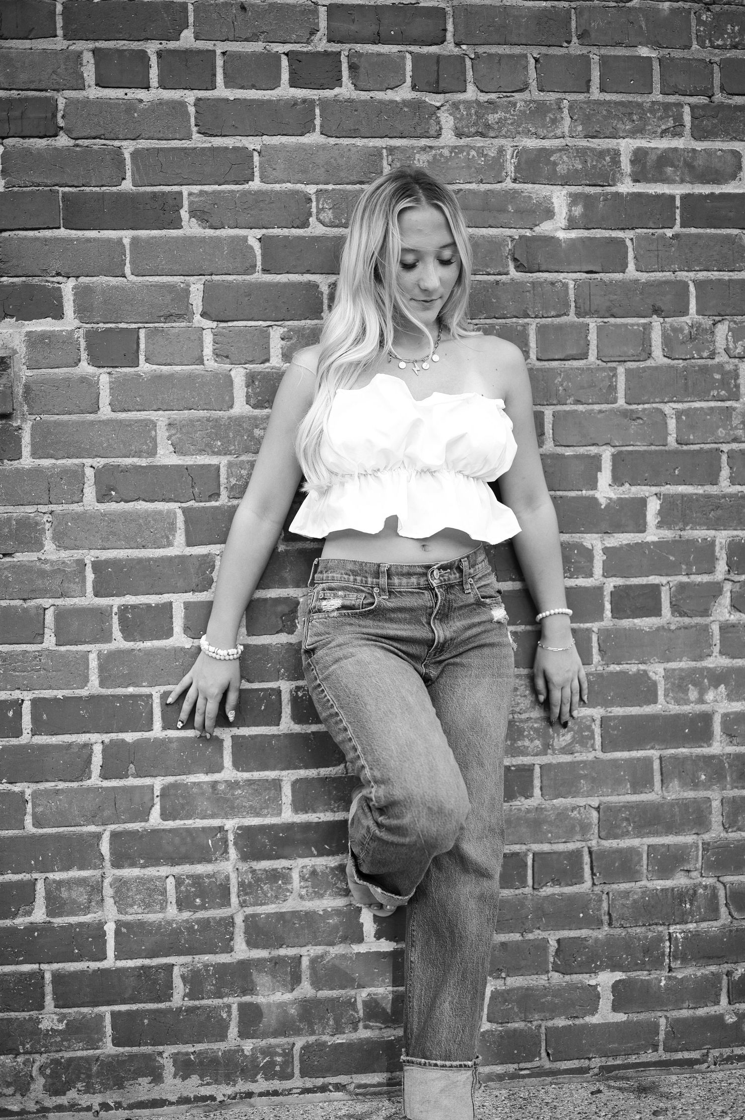 Black and white portrait of a young woman with long blonde hair leaning against a brick wall in a ruffled crop top and cuffed jeans.