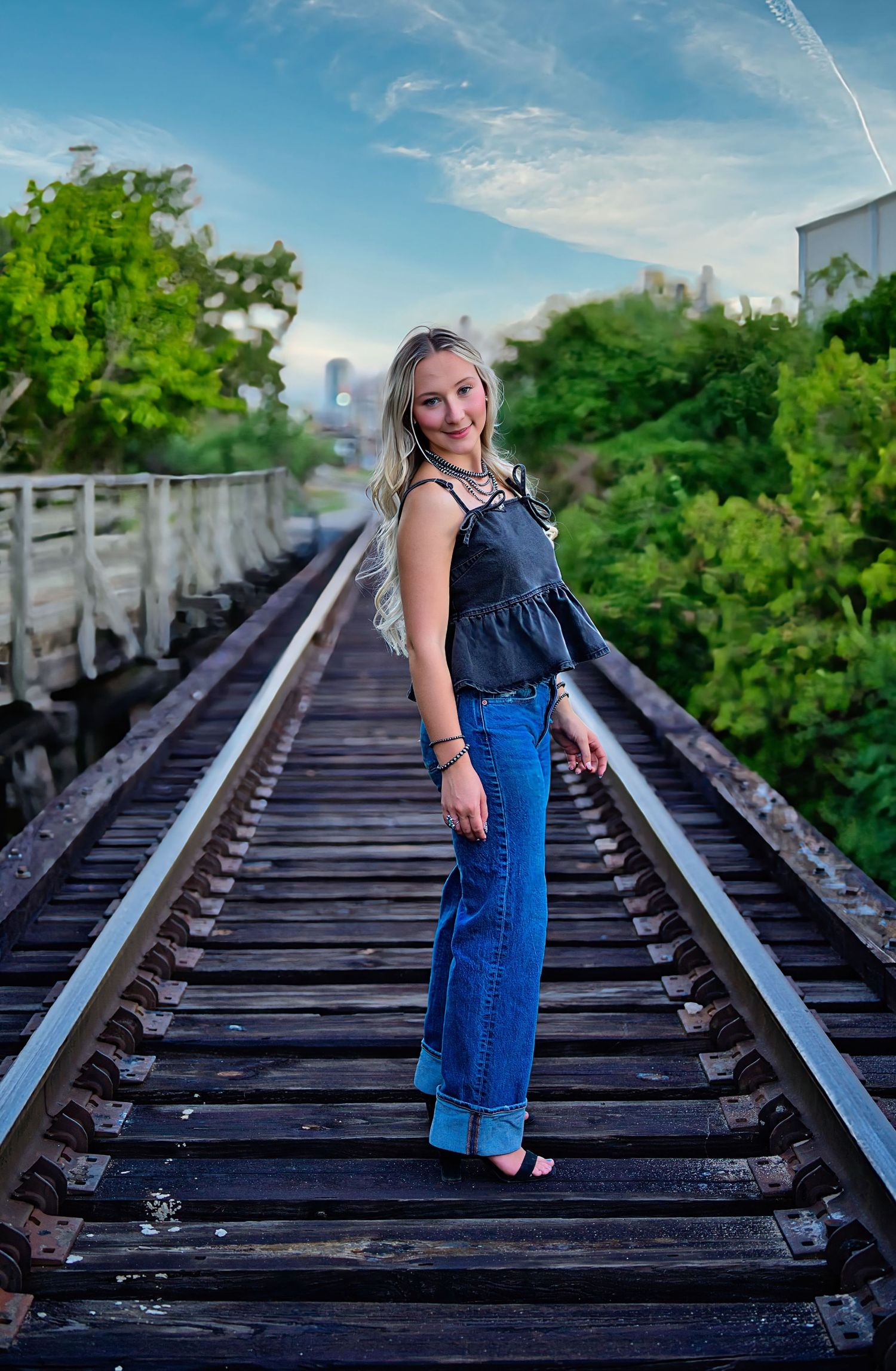 Young woman with long blonde hair poses on railroad tracks in a navy ruffled top and blue jeans, with green trees and a city skyline behind.