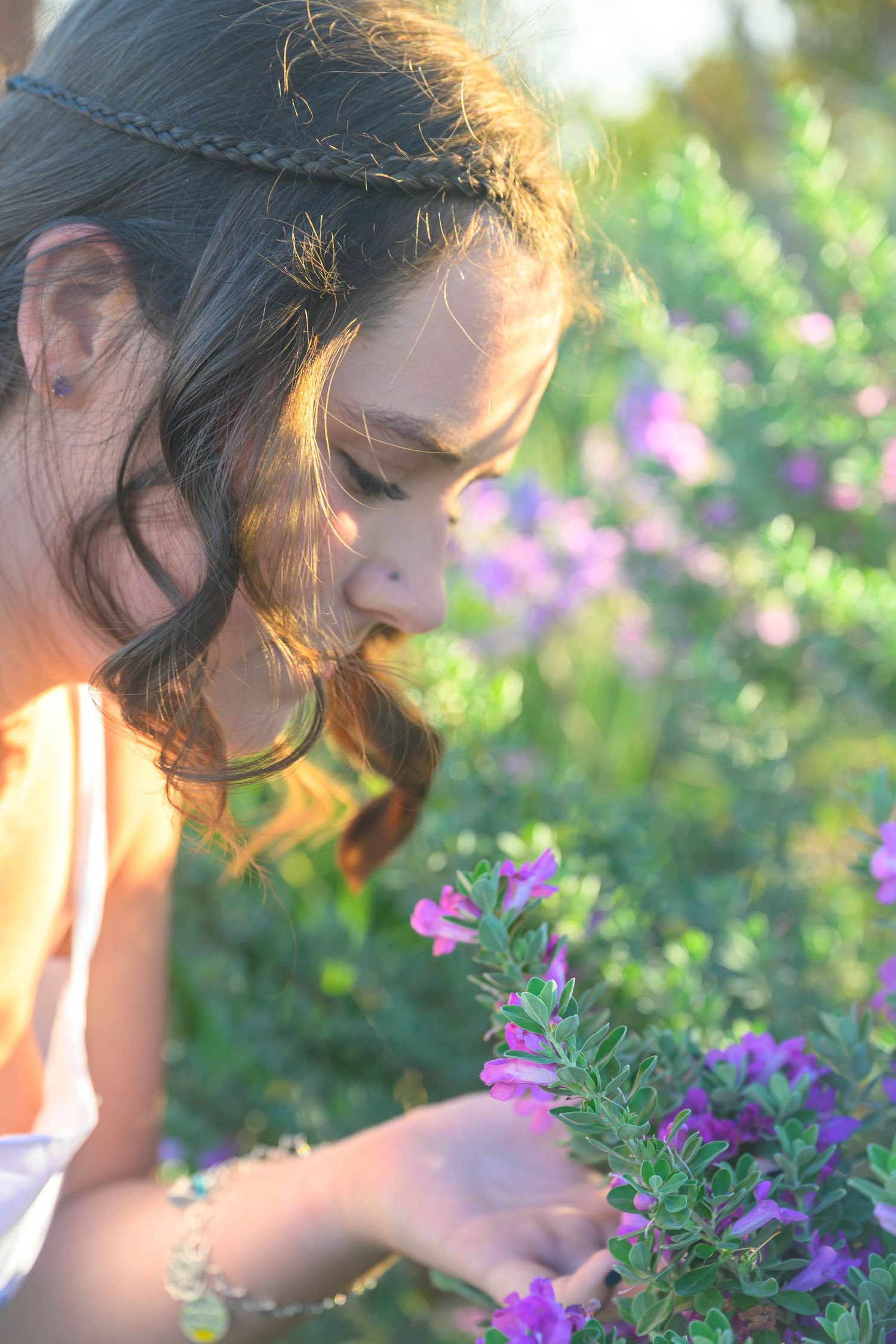 Young woman with a braided headband and wavy dark hair leans in to smell purple wildflowers in a sunlit garden.
