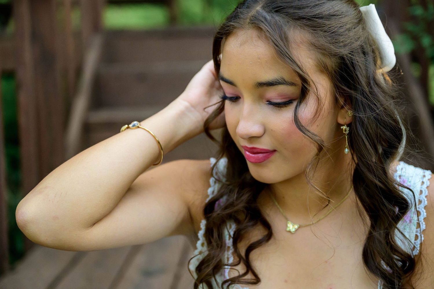 Young woman with curly hair wearing gold butterfly jewelry and a white bow, posing outdoors near wooden stairs.