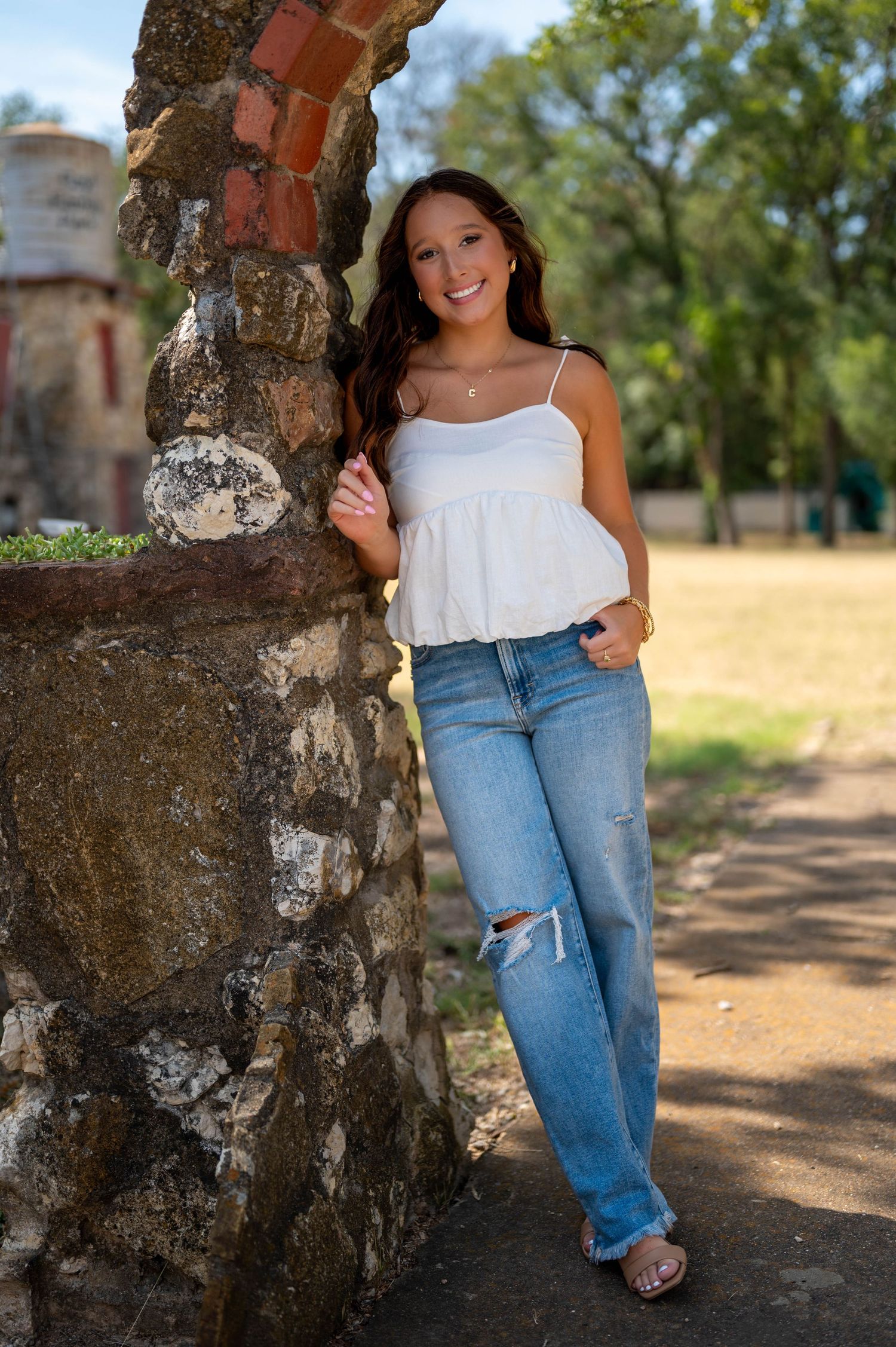 A casual portrait session against a rustic stone wall showcases relaxed poses in blue jeans and a white top. Azle senior at the park.