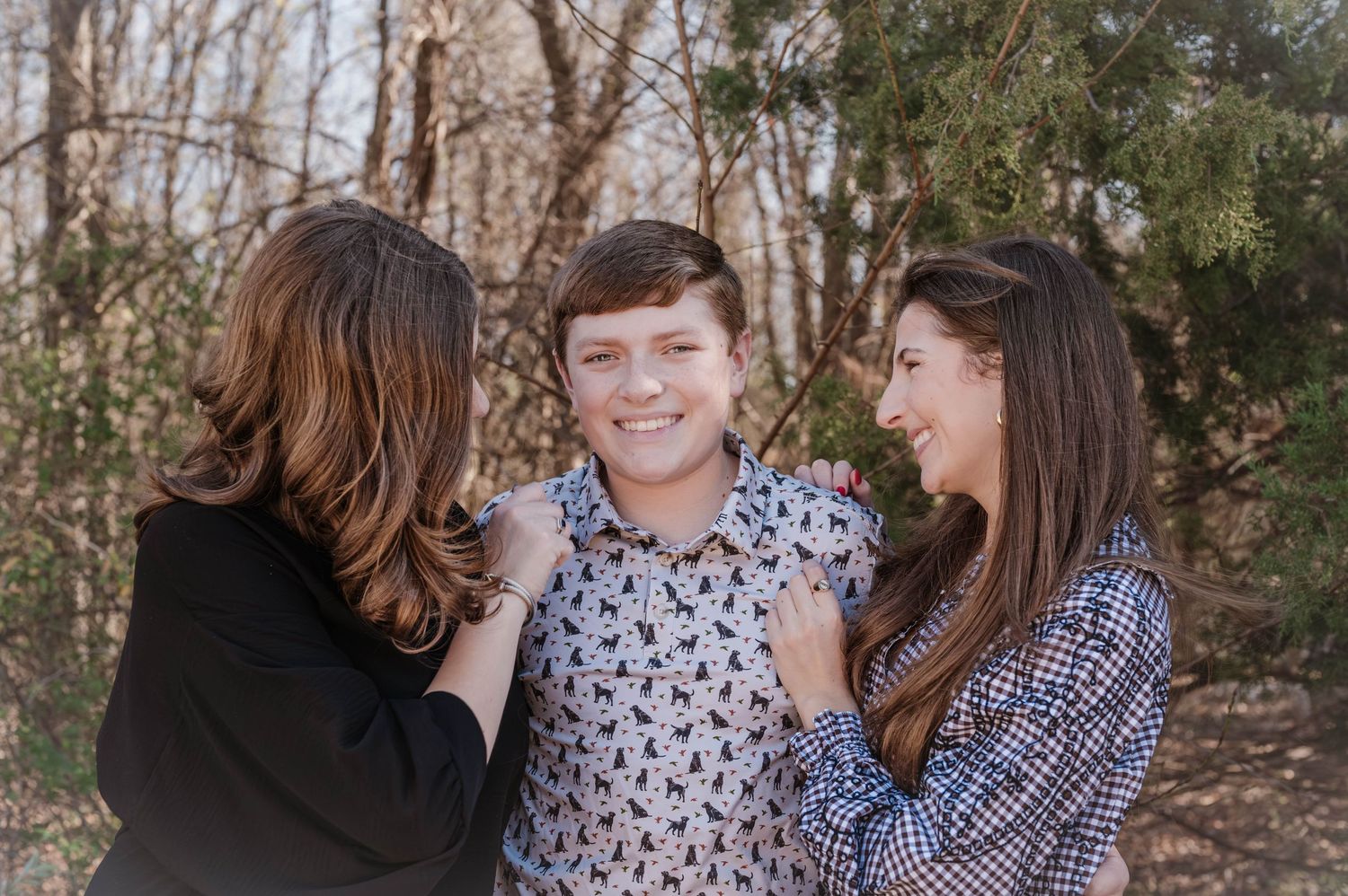Smiling teenage boy in a patterned button-up shirt stands between two women who lean in affectionately during an outdoor family portrait.