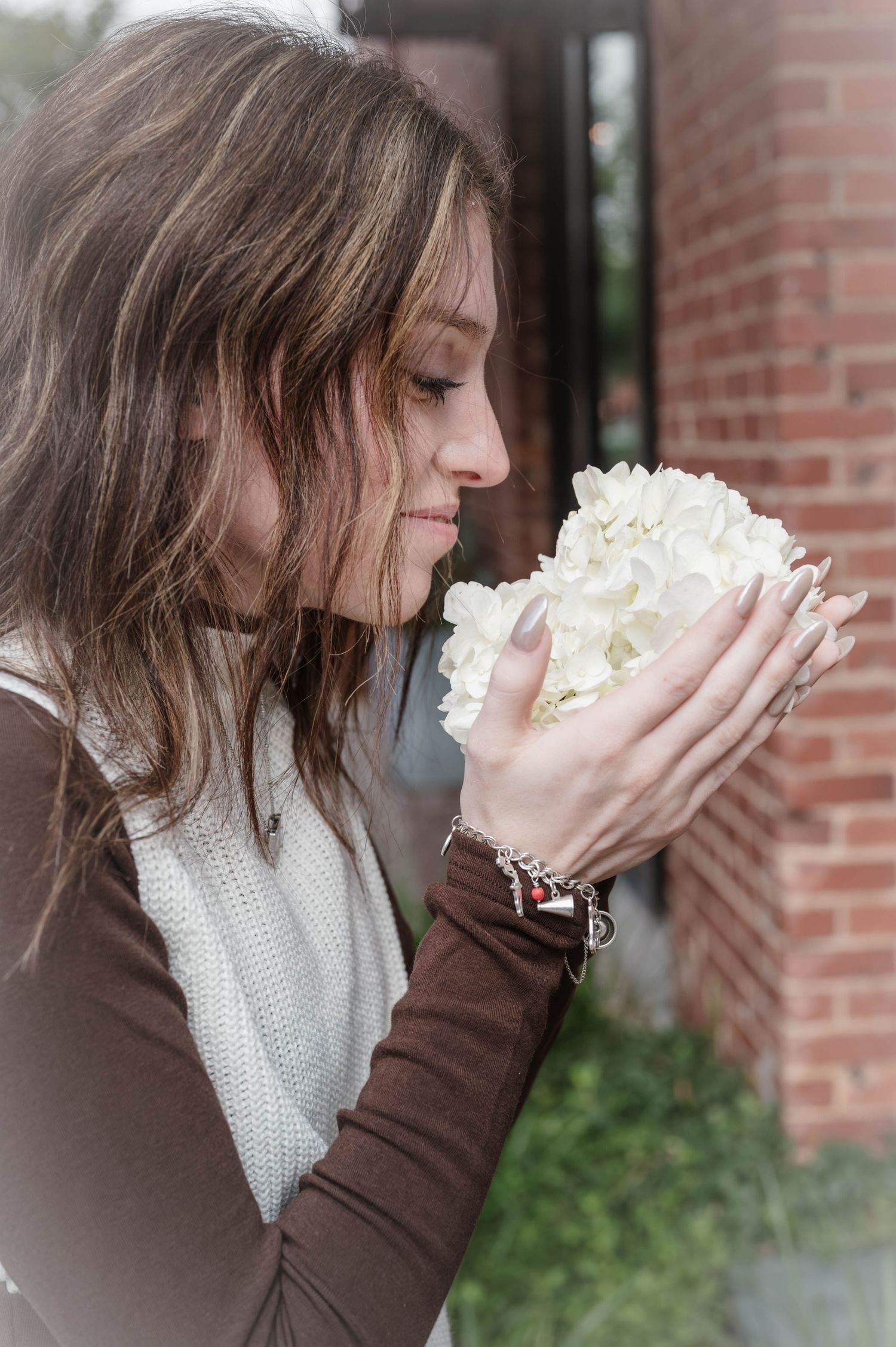 Woman with long brown hair in a brown top and cream scarf gently smells a white hydrangea bouquet beside a red brick wall.
