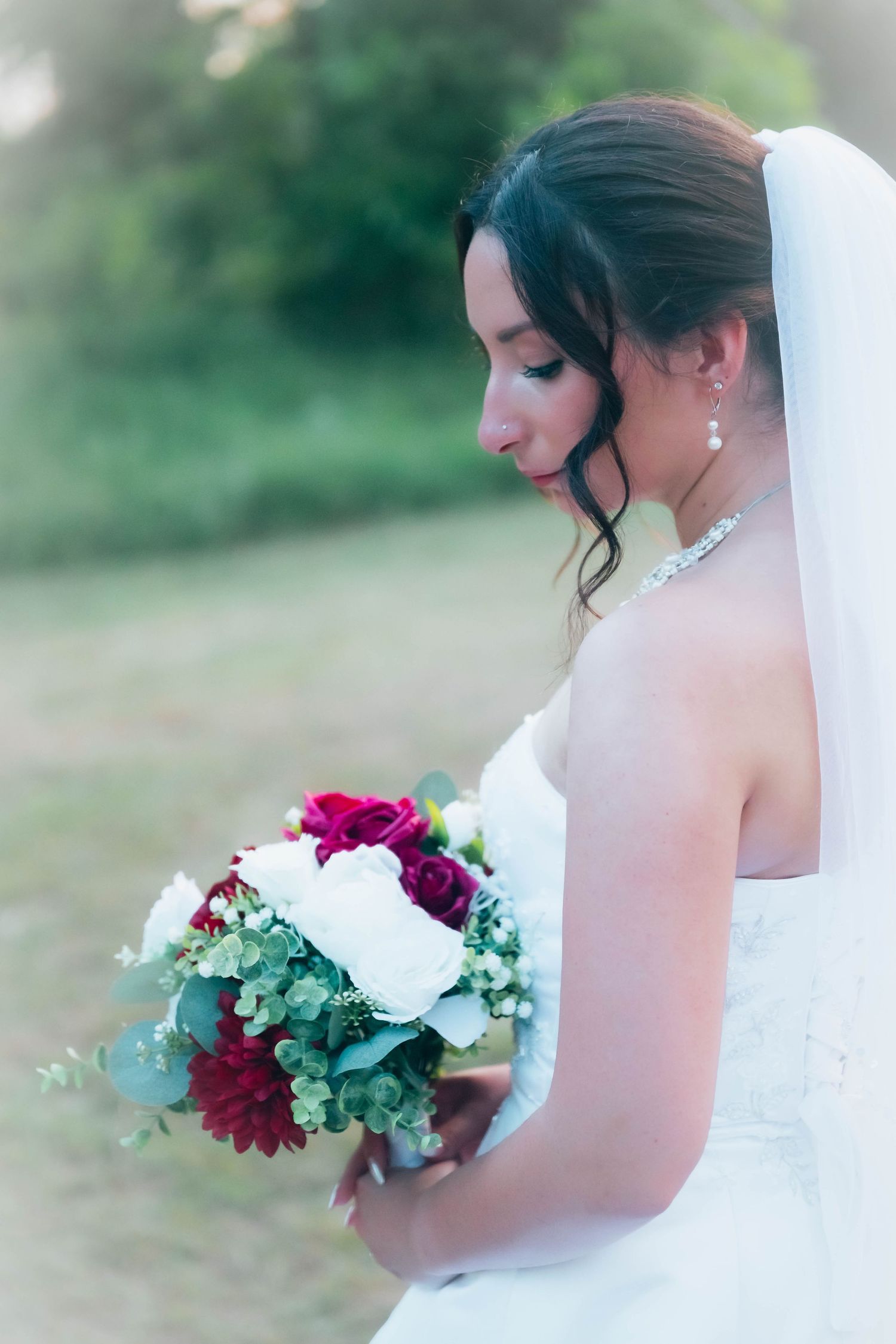 Bride in white gown holding red and white floral bouquet with greenery, looking down gracefully outdoors.