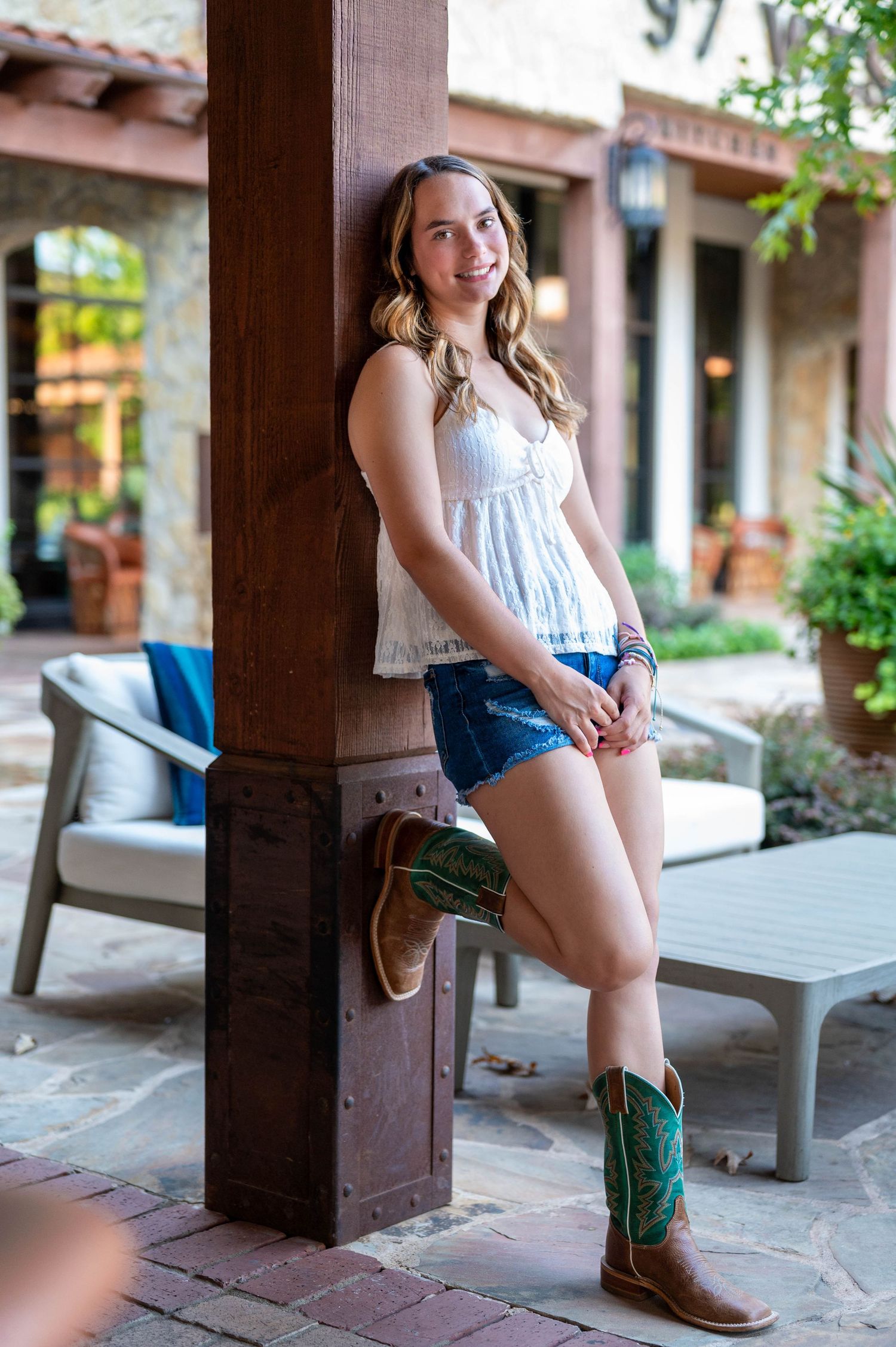 A girl wearing blue denim shorts and cowboy boots sits against a column under a covered porch. Azle senior at the fort worth stockyards.