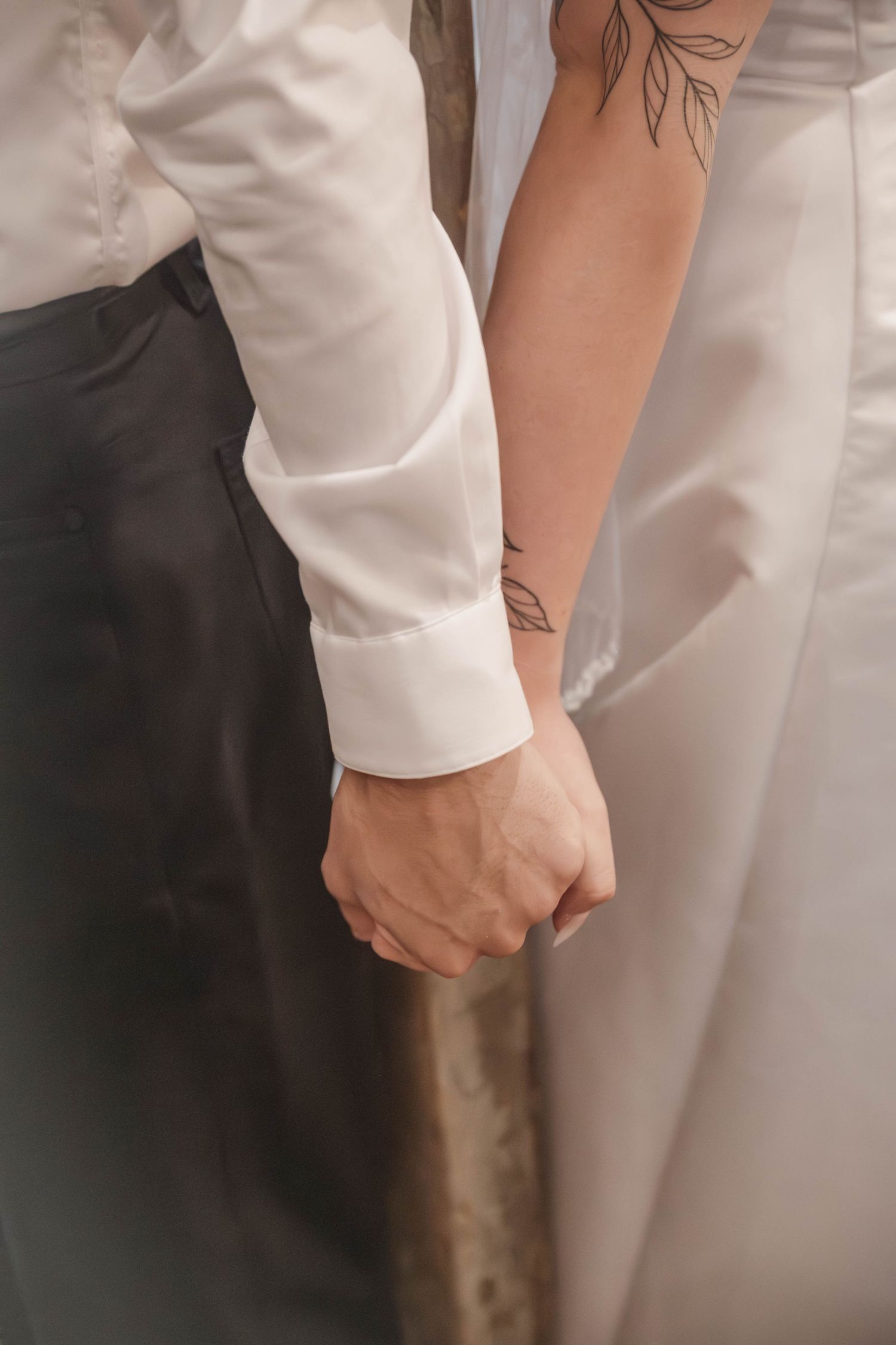 Close-up of a bride and groom holding hands, showing her floral wrist tattoo, white dress, and his white shirt.