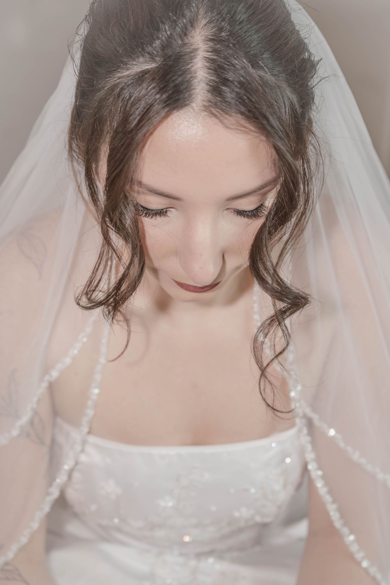 Bride in white beaded gown and pearl-trimmed veil looking down, soft romantic bridal portrait photography.