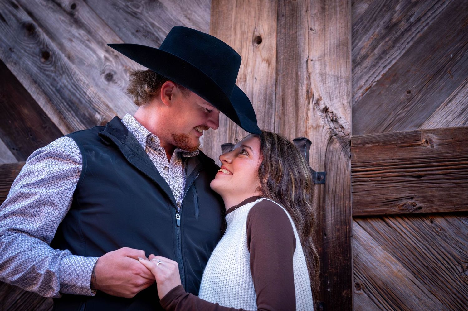 Smiling couple in western attire hold hands and share a tender moment in front of a rustic weathered barn wood wall.