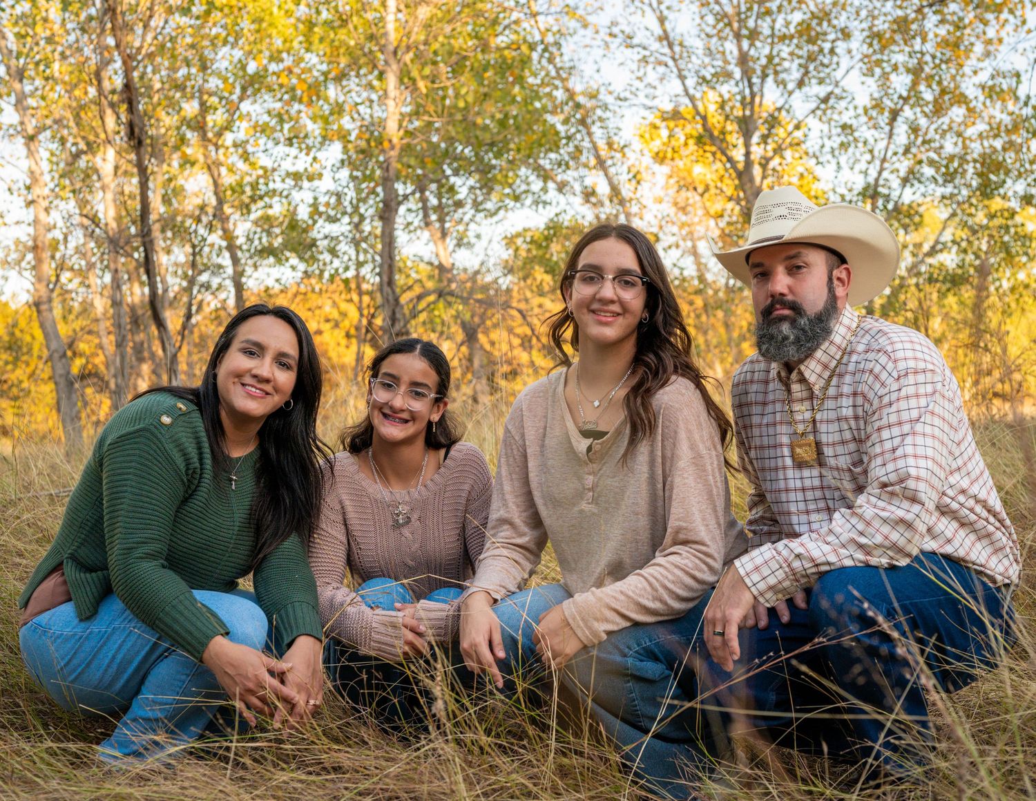 Four people, including a bearded man in a cowboy hat, sit close together in a grassy field with golden autumn trees in the background.