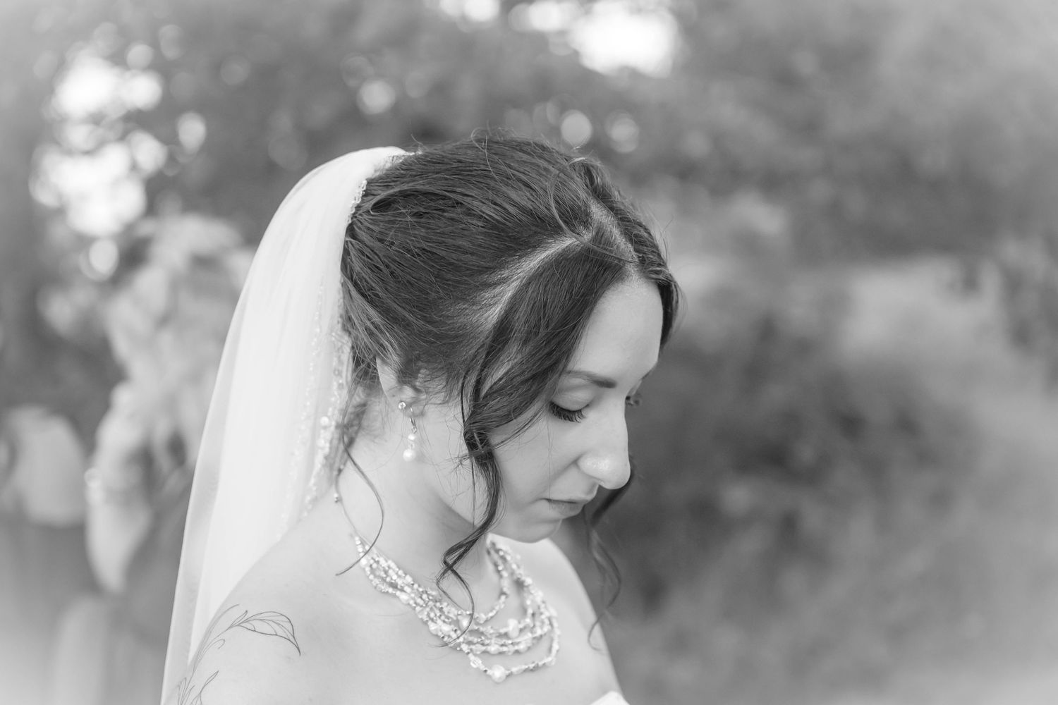 Black and white bridal portrait of a bride in veil and pearl necklace looking down outdoors.