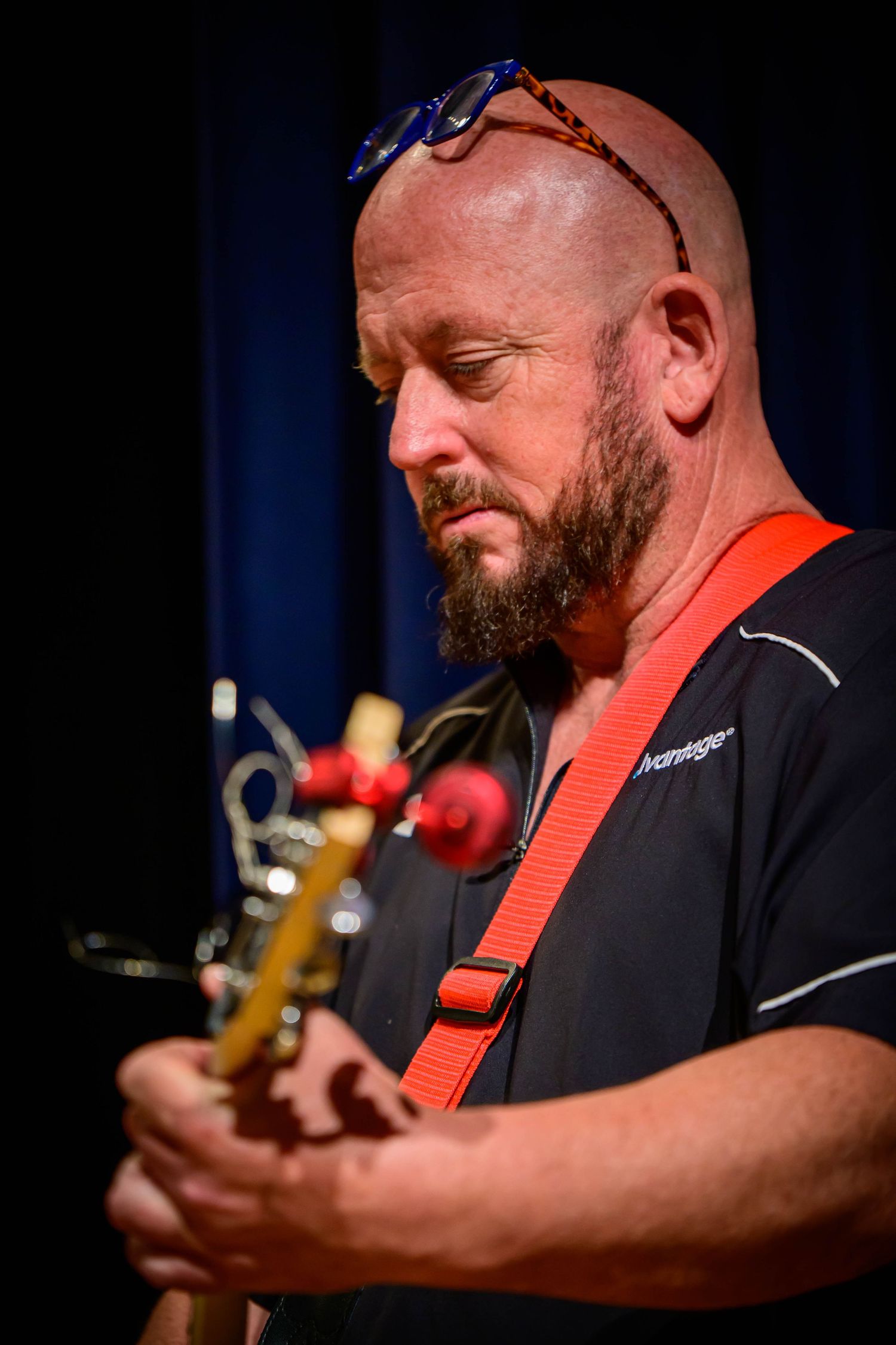 Bald bearded man with sunglasses on his head looks down while playing a guitar with a red strap during a live performance.
