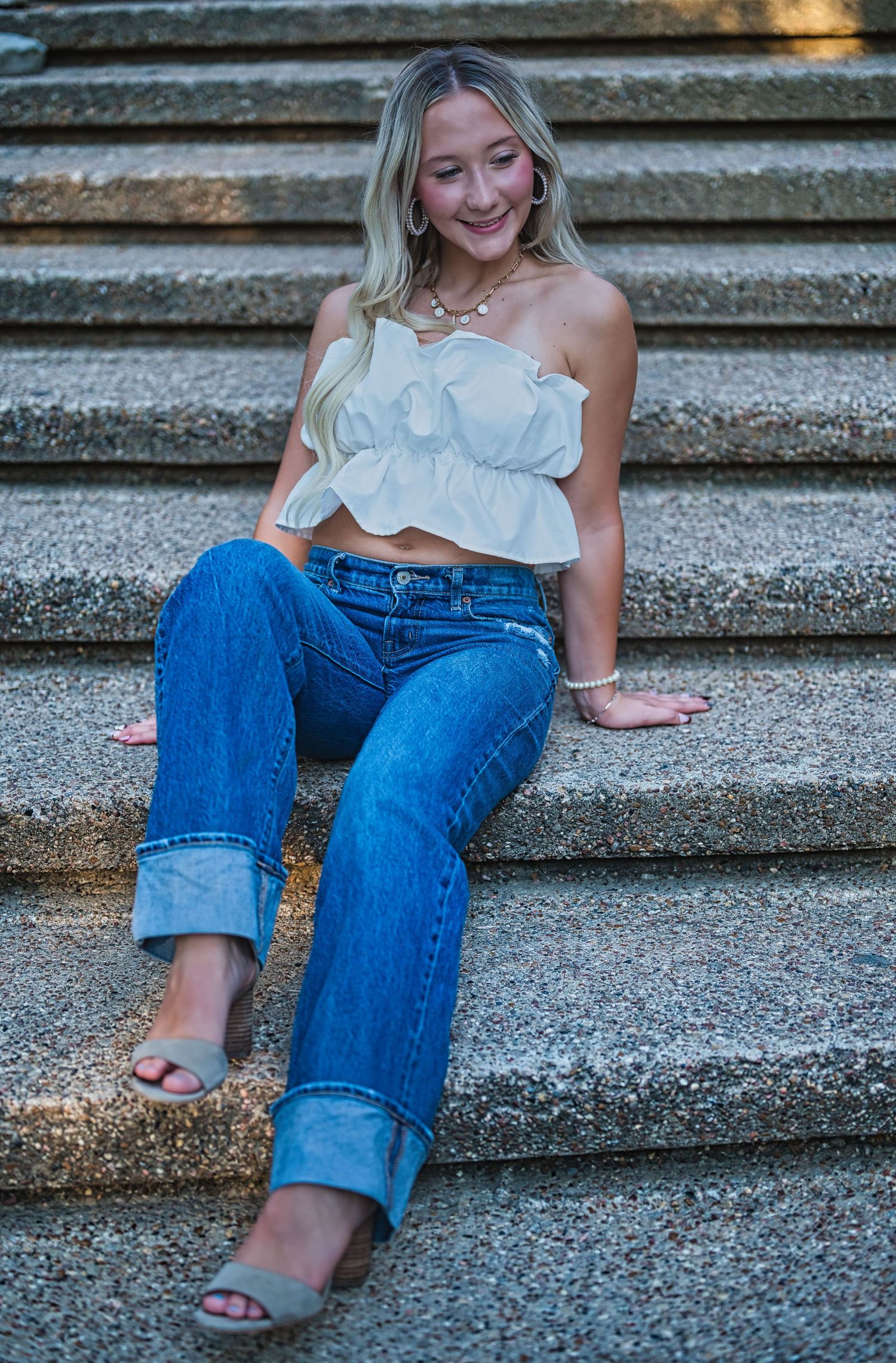 Smiling young woman with long blonde hair sits on stone steps wearing a white ruffled crop top, cuffed blue jeans and gray heeled sandals.