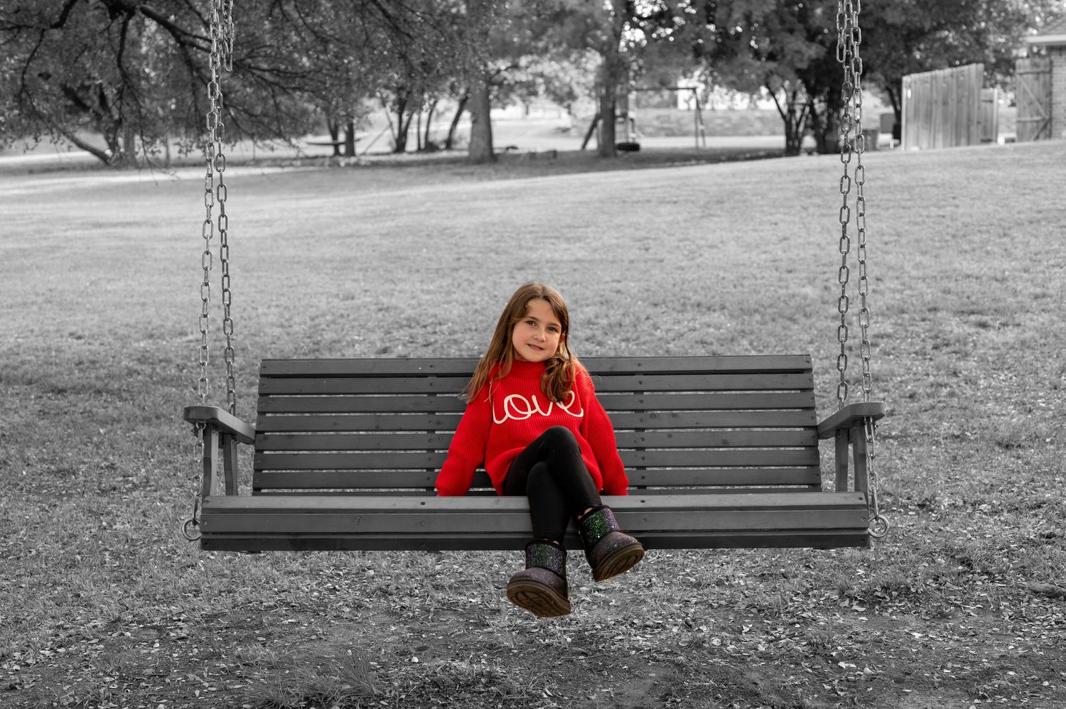 A wooden swing bench in a park with a little girl in a bright red sweater against black and white background. Azle family photo session.