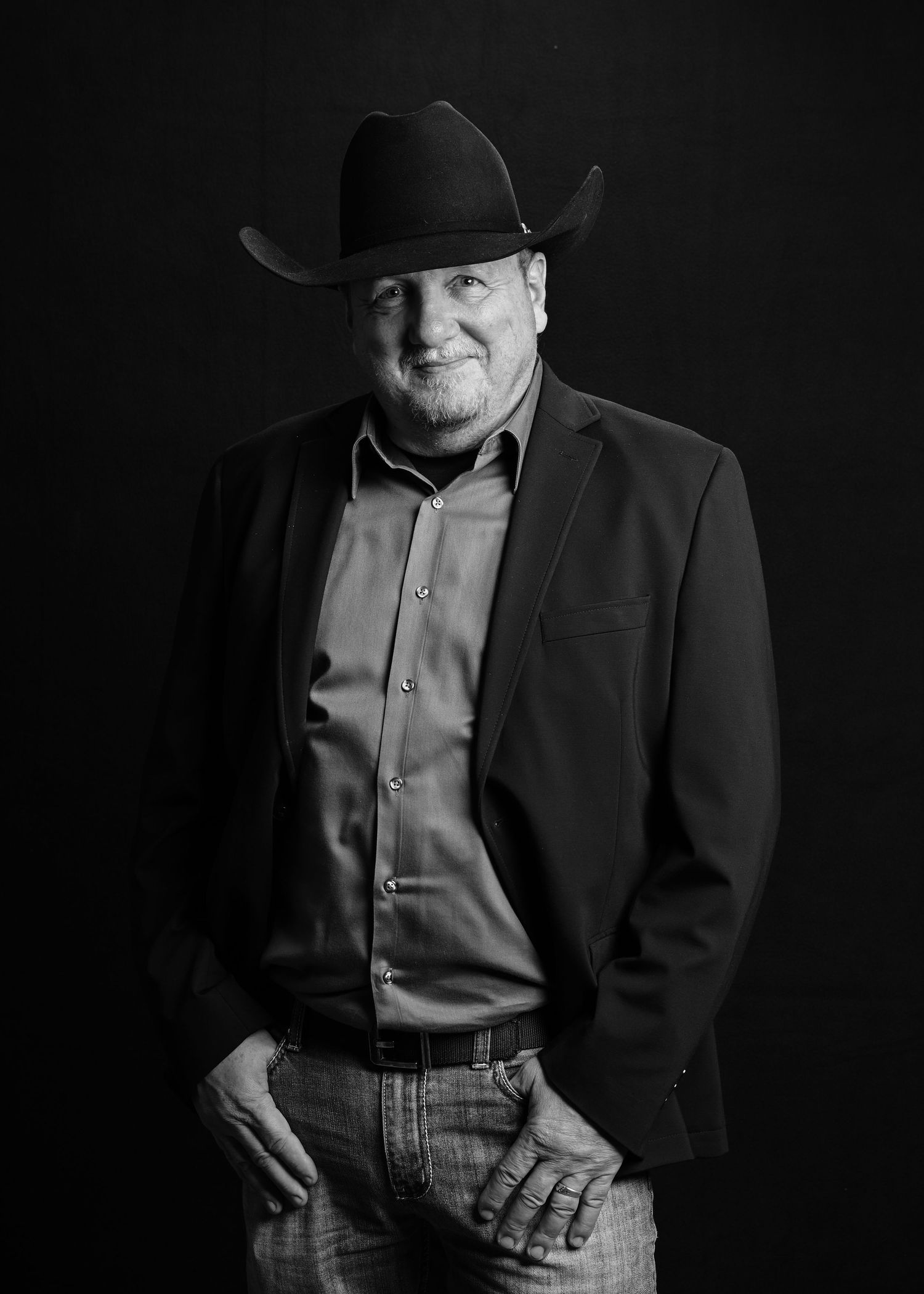 Black and white portrait of a person wearing a cowboy hat, suit jacket and button-up shirt against dark background.