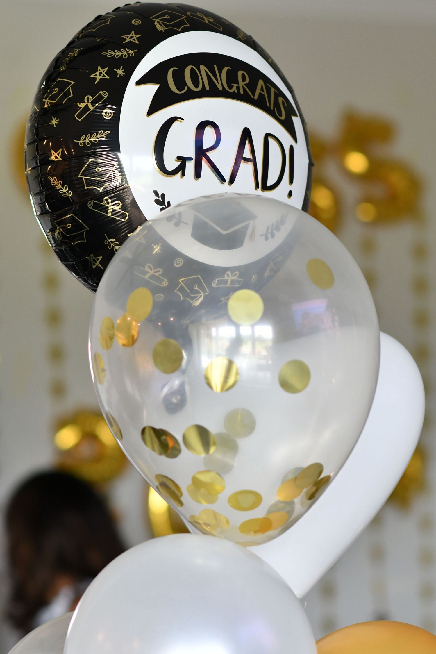 Clear graduation balloons decorated with gold confetti dots and 'GRAD!' text float against a blurred background.