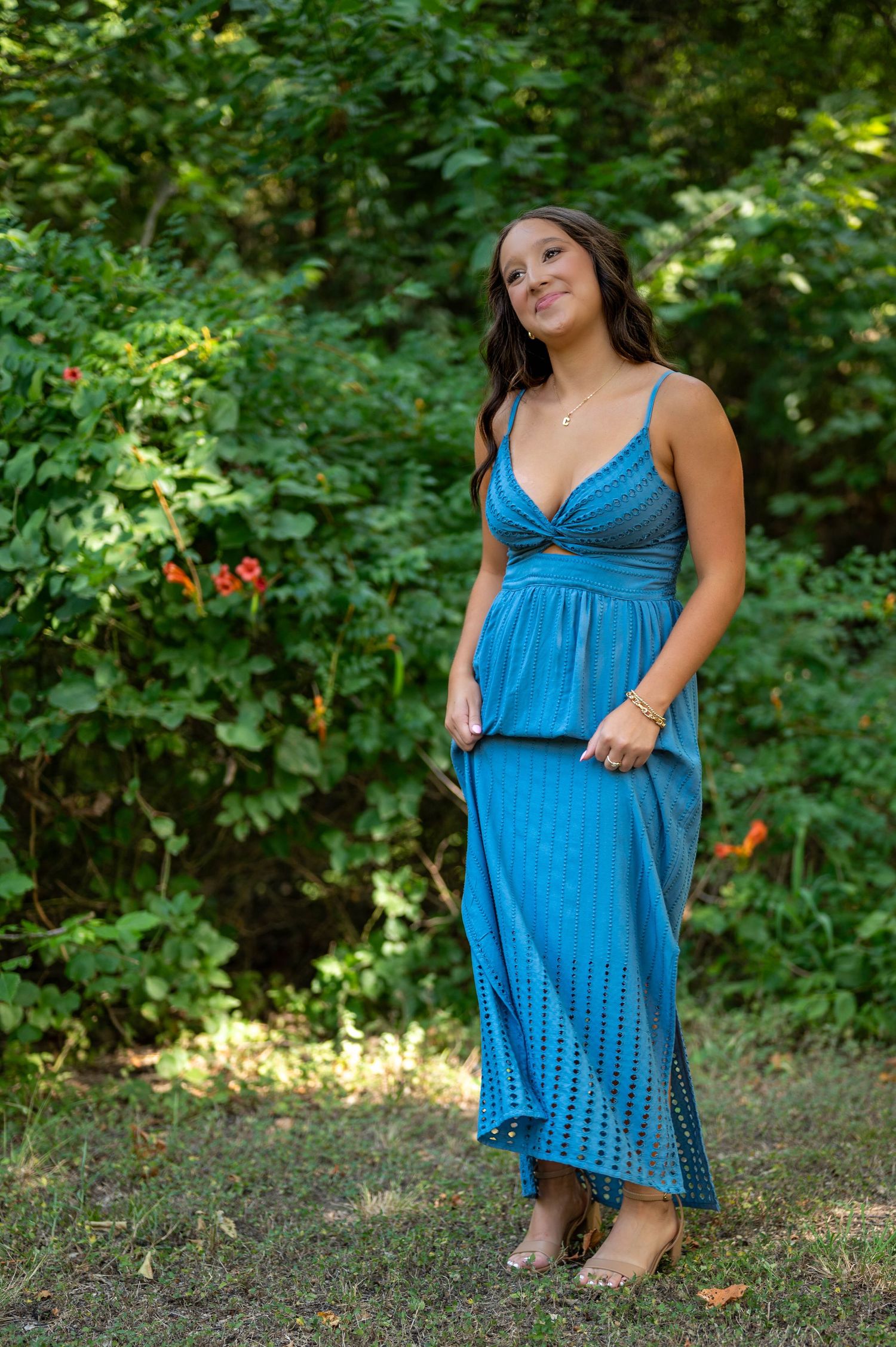 A person in a flowing blue summer dress poses outdoors against lush green foliage in natural lighting. Azle senior at the park.