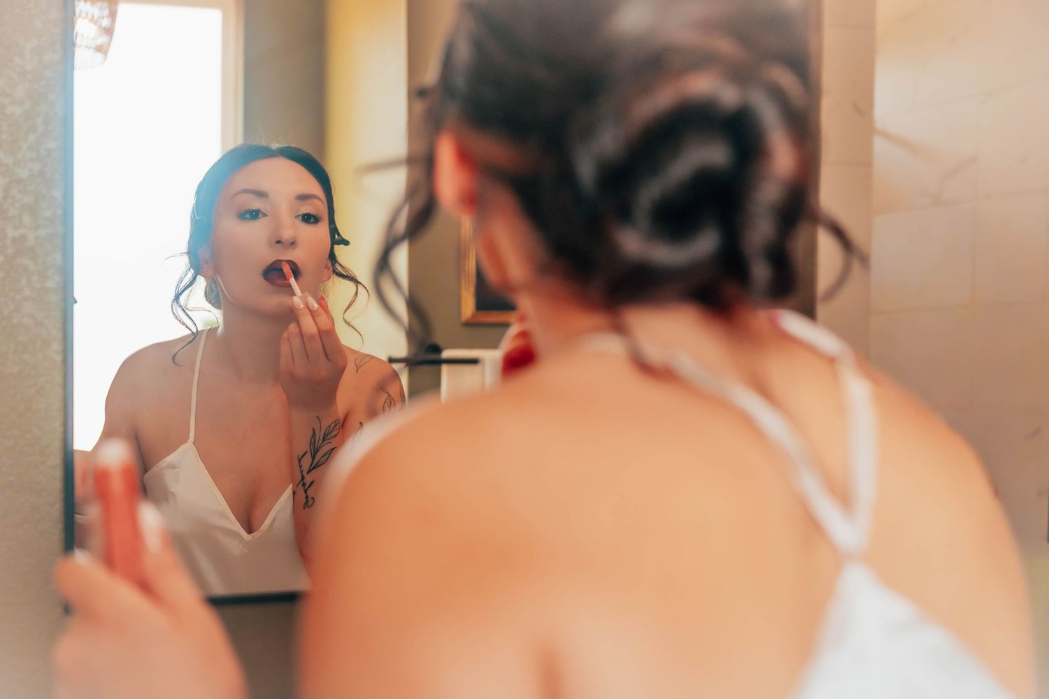 Woman in a white strappy top applies red lipstick while looking at her reflection in a mirror, her hair pinned up in a loose bun.