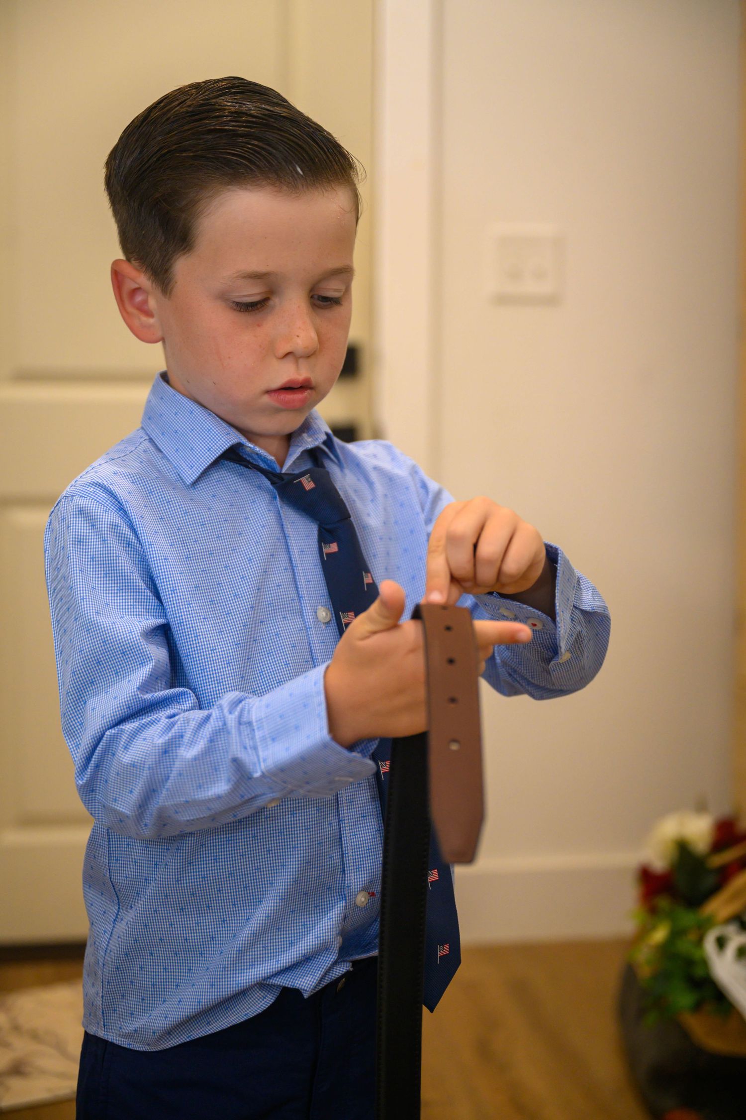 A young boy in a blue button-up shirt and patterned tie looks down while threading a brown leather belt indoors.
