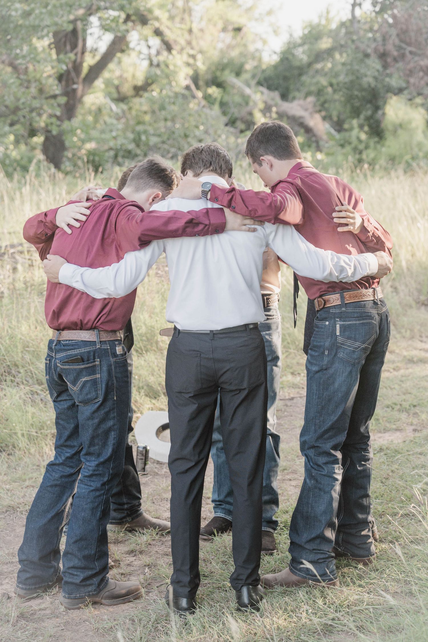 Four young men in matching maroon shirts and jeans huddle together in a group embrace outdoors in a field.