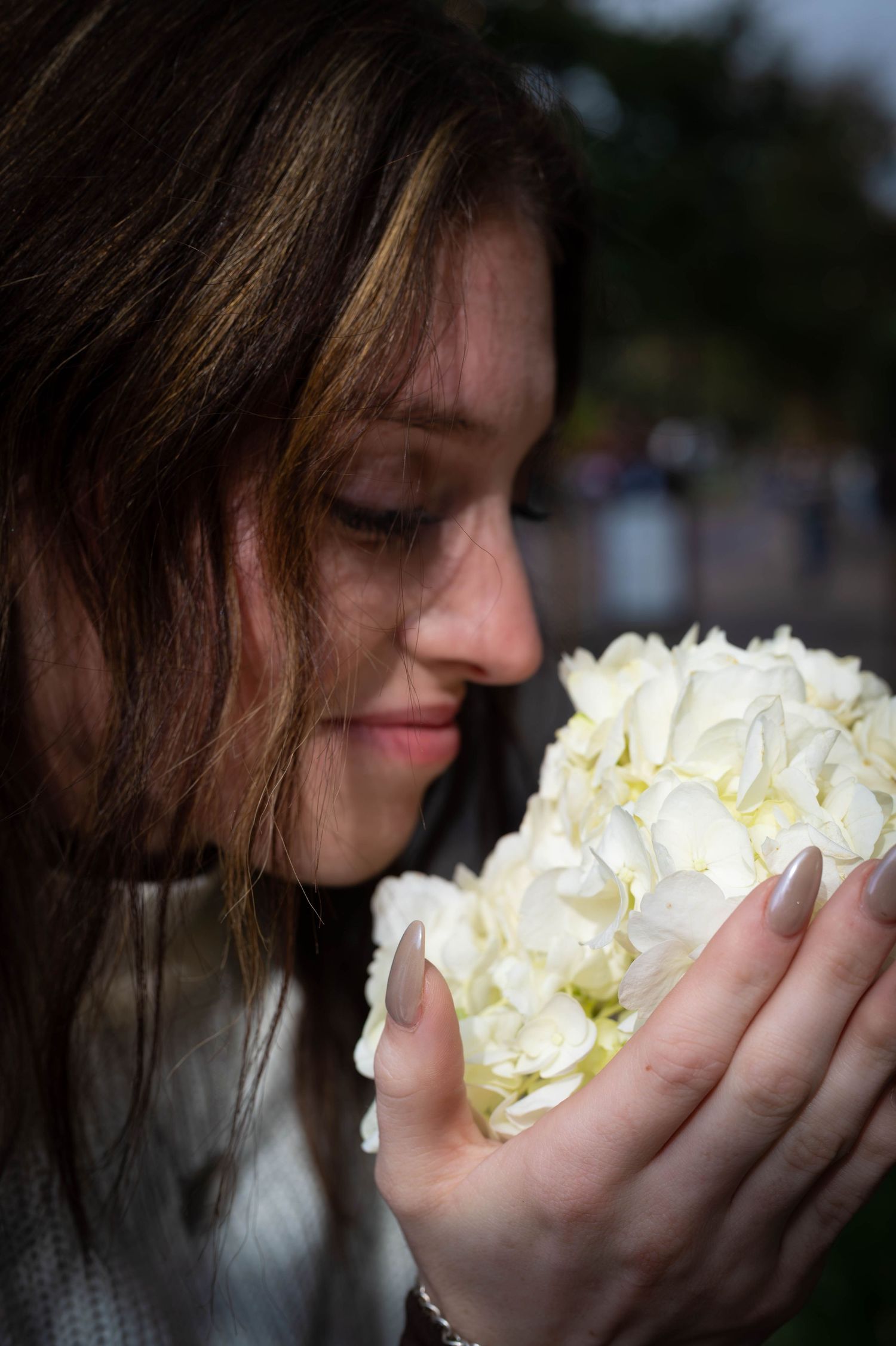 Young woman with long brown hair smelling a bouquet of white hydrangea flowers, eyes closed in enjoyment.