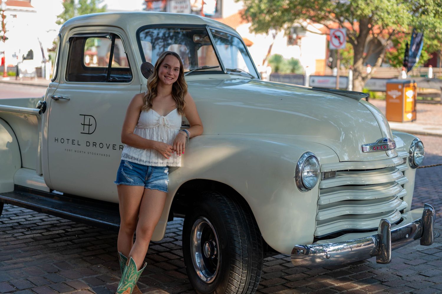 Smiling woman in denim shorts and green cowboy boots leans on a vintage mint green Chevrolet truck with Hotel Drover logo on a brick street.