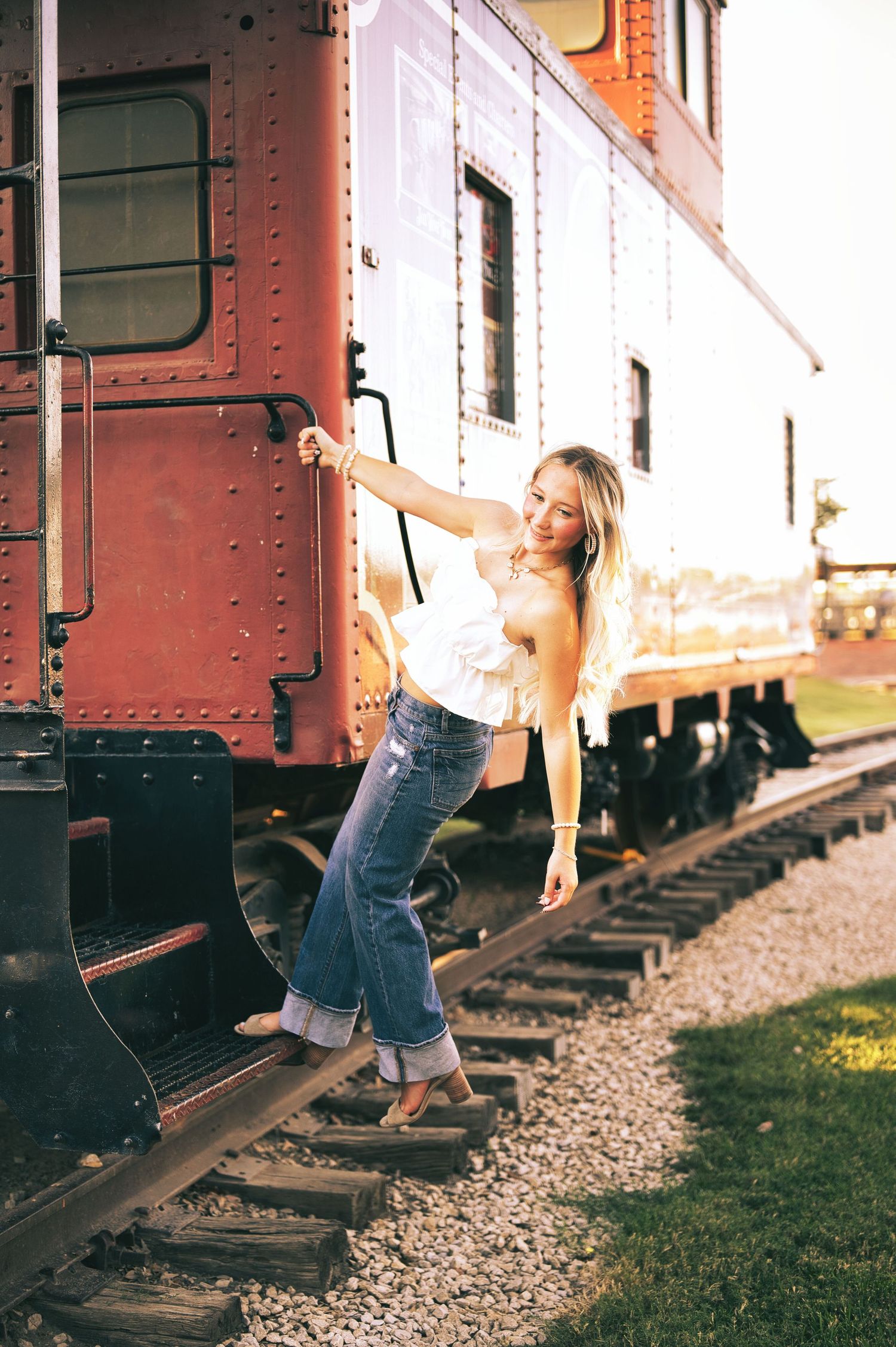 Young woman in a white ruffled top and jeans leans off the step of a vintage red caboose on railroad tracks, smiling in warm sunlight.