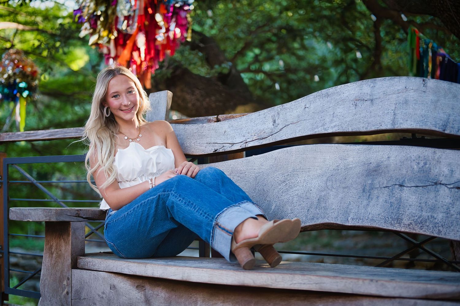 A person in a white off-shoulder top and blue jeans relaxes on a curved wooden bench outdoors. Azle senior at the fort worth stockyards.