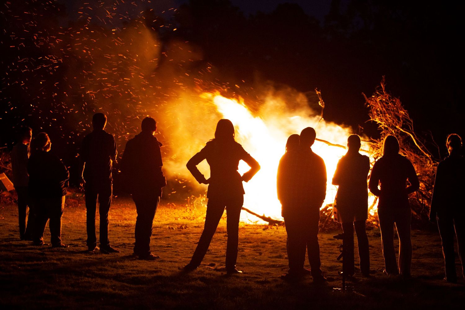 Silhouetted people stand in a semicircle around a large bonfire at night, with bright orange flames and sparks lighting the dark sky.