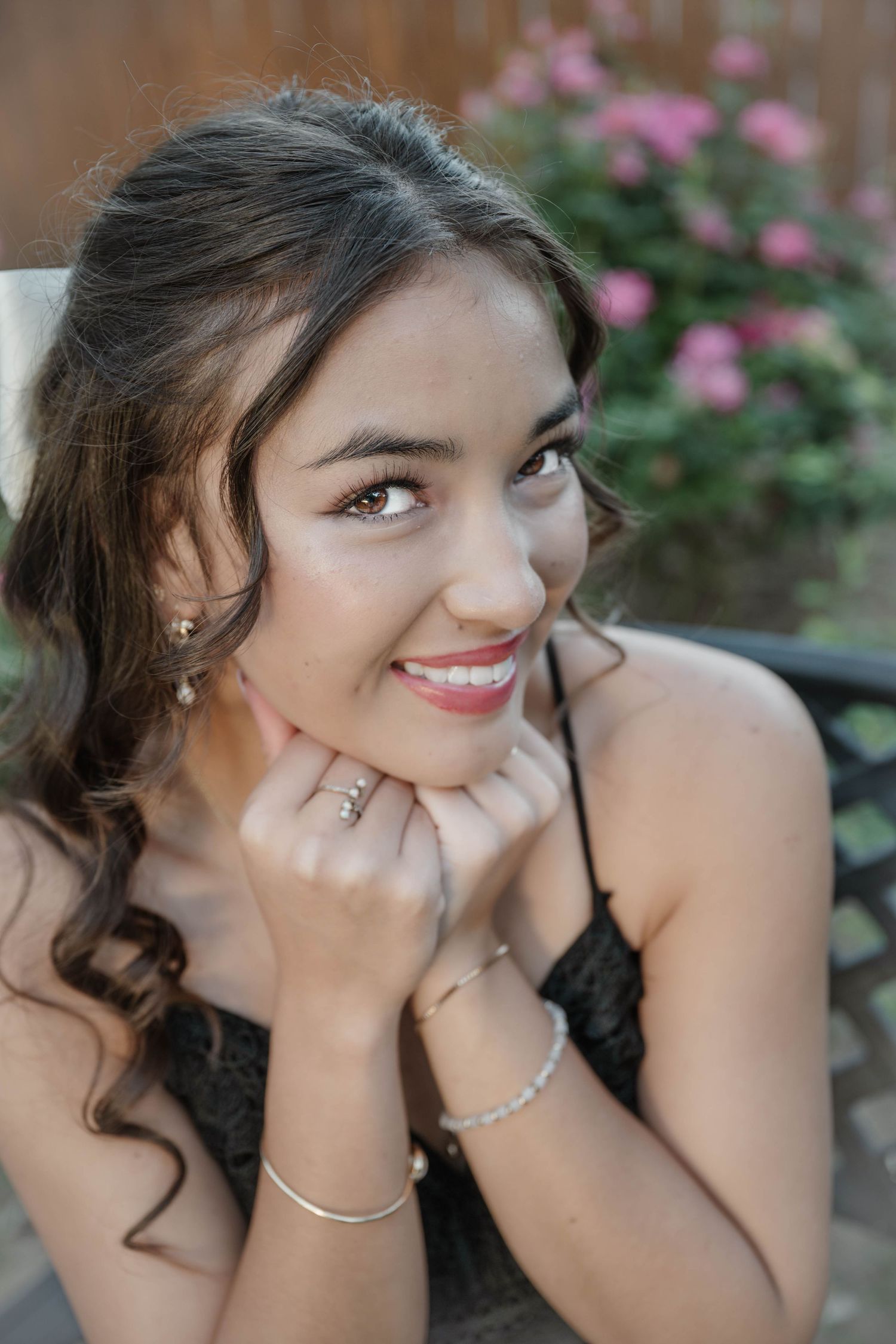 Smiling young woman with curly hair, wearing a black dress, silver bracelet, and rings, posing outdoors near pink flowers.