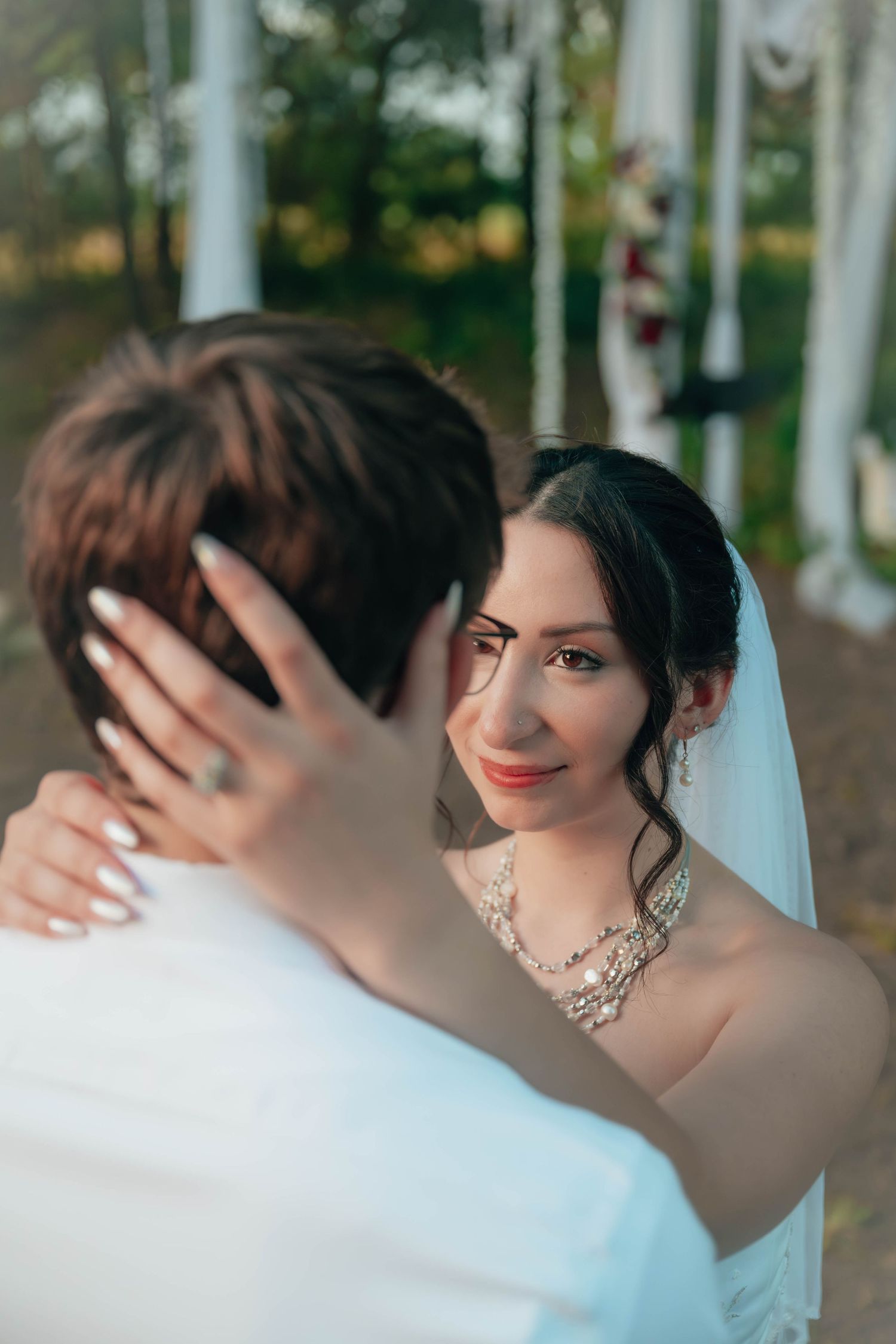 Bride in white veil and necklace gazes lovingly at groom during outdoor wedding ceremony.