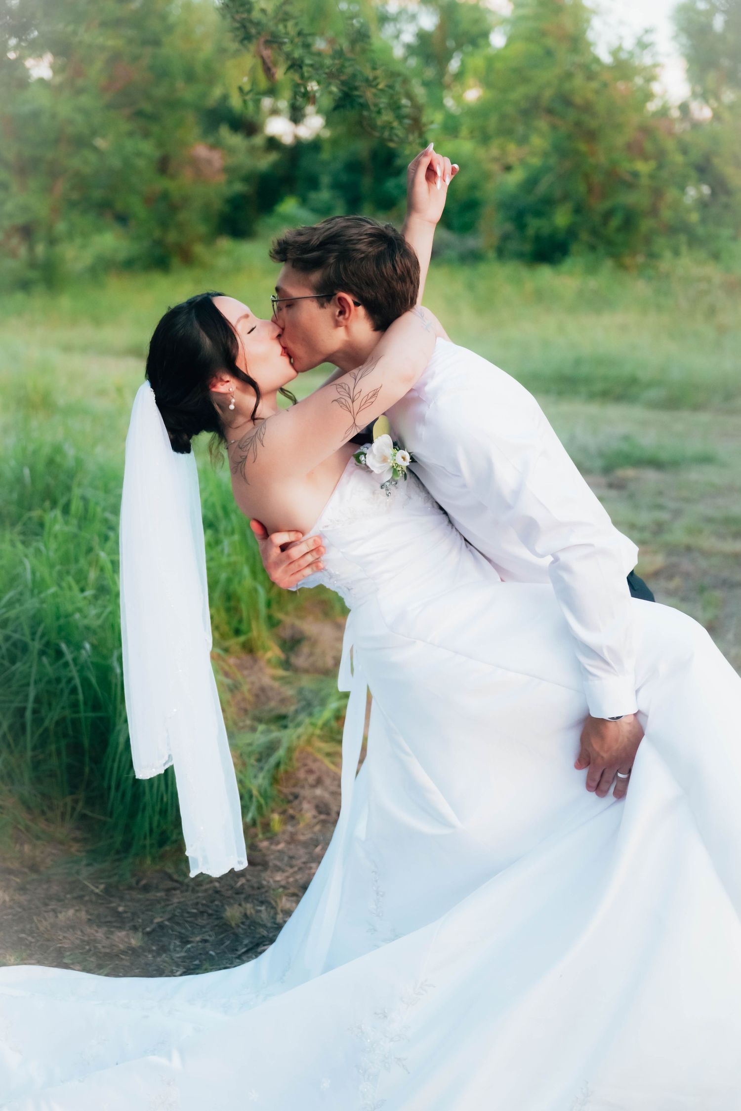 A groom dips his tattooed bride for a romantic kiss outdoors, her white wedding dress and veil flowing gracefully.