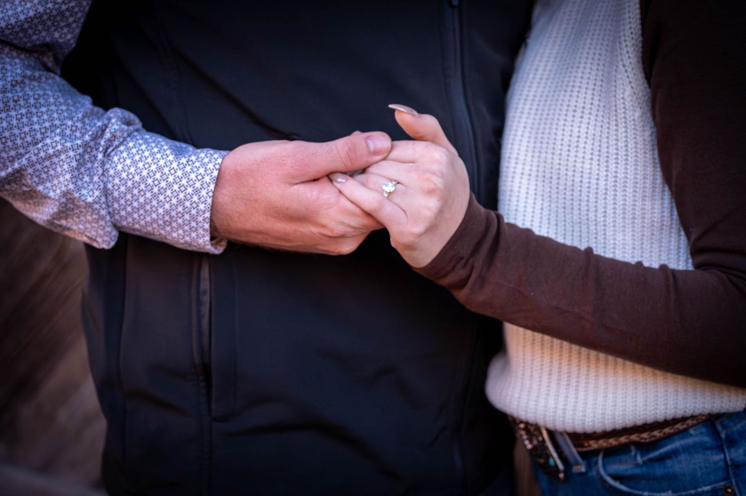 Couple holding hands closely, showing a diamond engagement ring on her finger, in a black vest, patterned shirt and sweater.