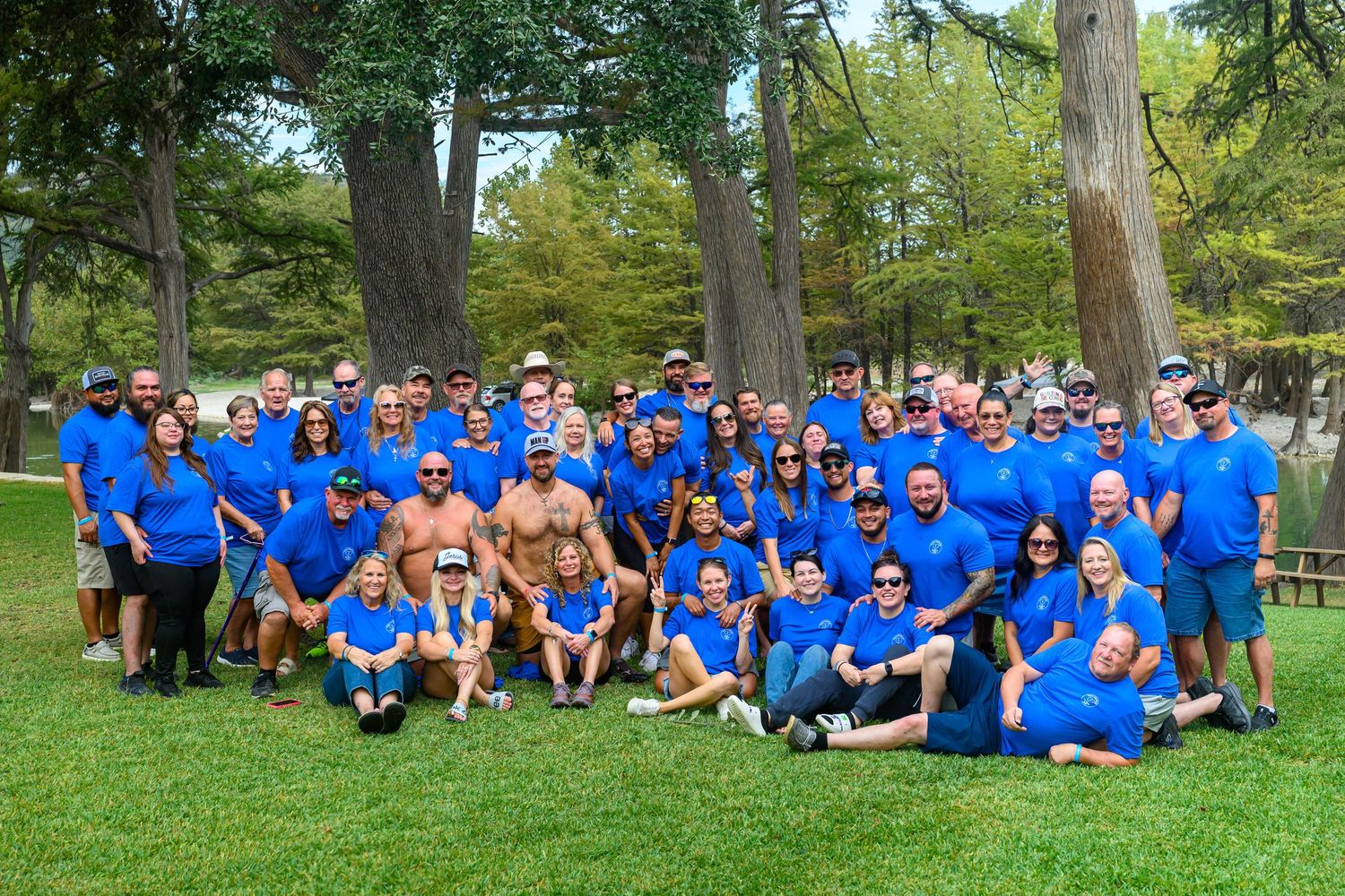 Large group of people in matching blue t-shirts posing together outdoors on a green lawn beneath tall trees on a sunny day.