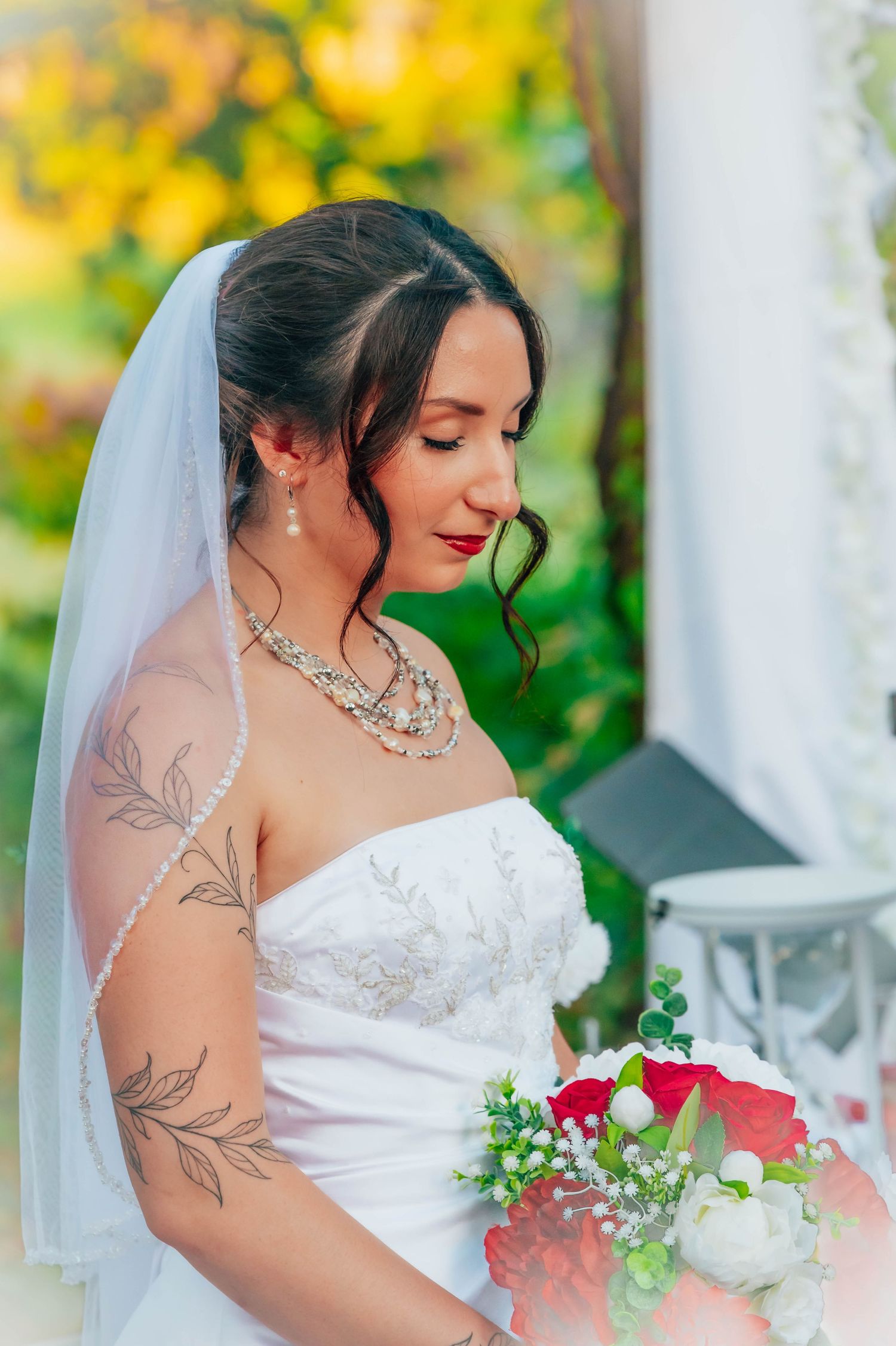 Bride in white strapless gown with veil and pearl necklace holds red and white bouquet, showcasing floral arm tattoo.