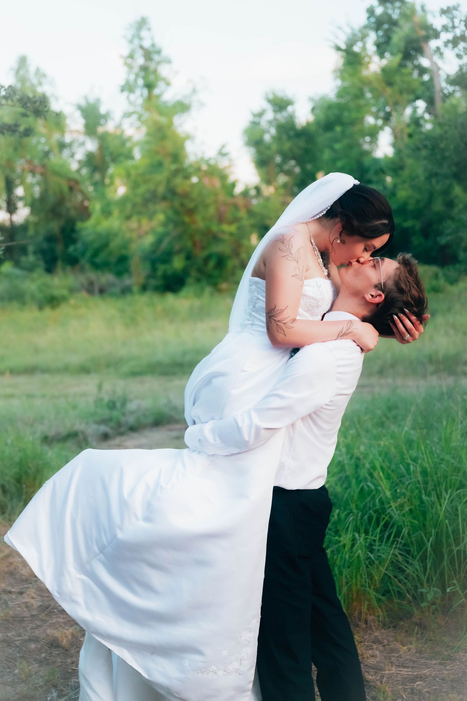 Groom lifting bride in white wedding dress and veil, sharing a romantic kiss outdoors in a lush green field.