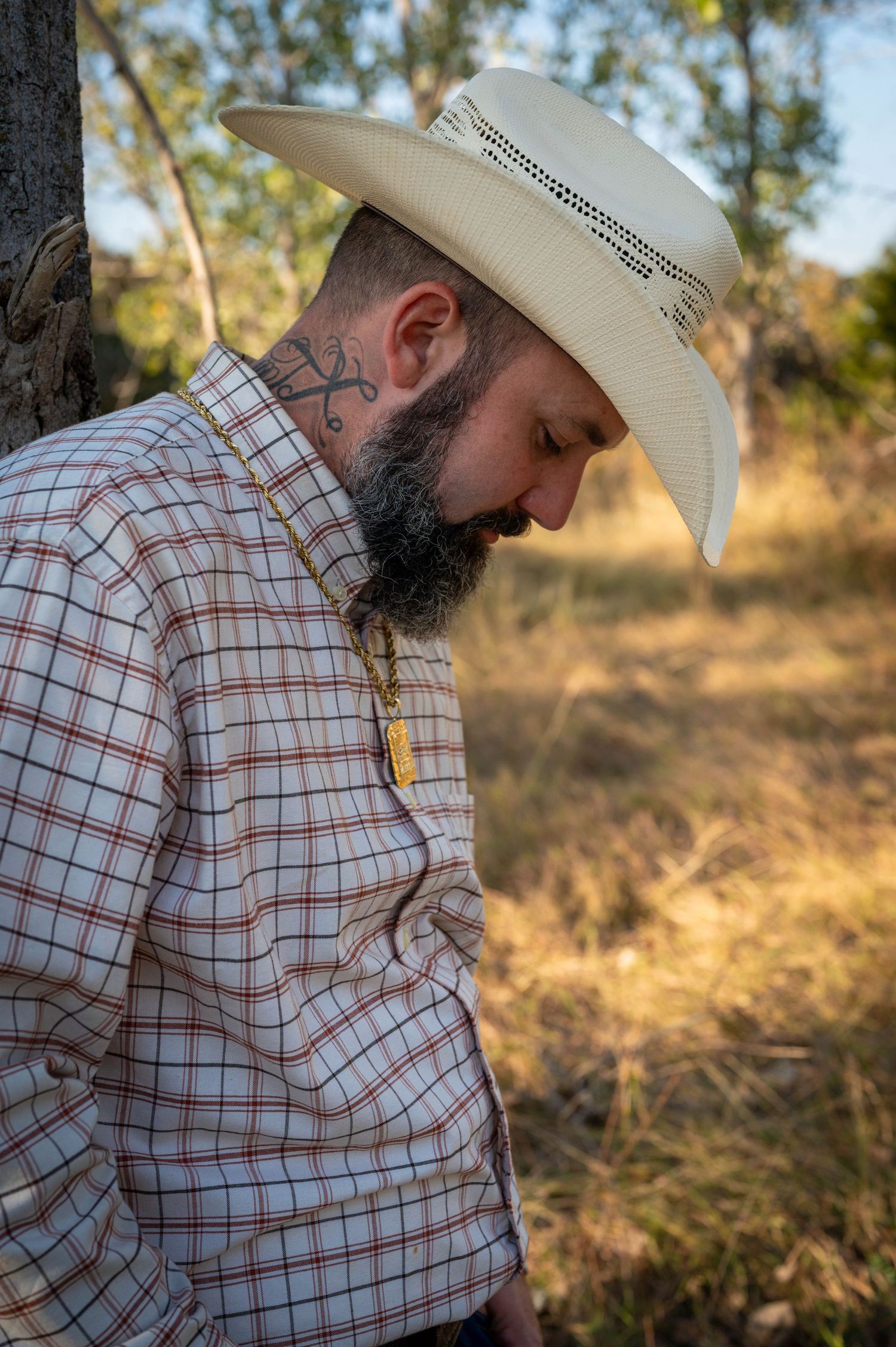 Bearded man in a cream straw cowboy hat and plaid western shirt looks down in a sunlit field, with a neck tattoo and gold pendant necklace.