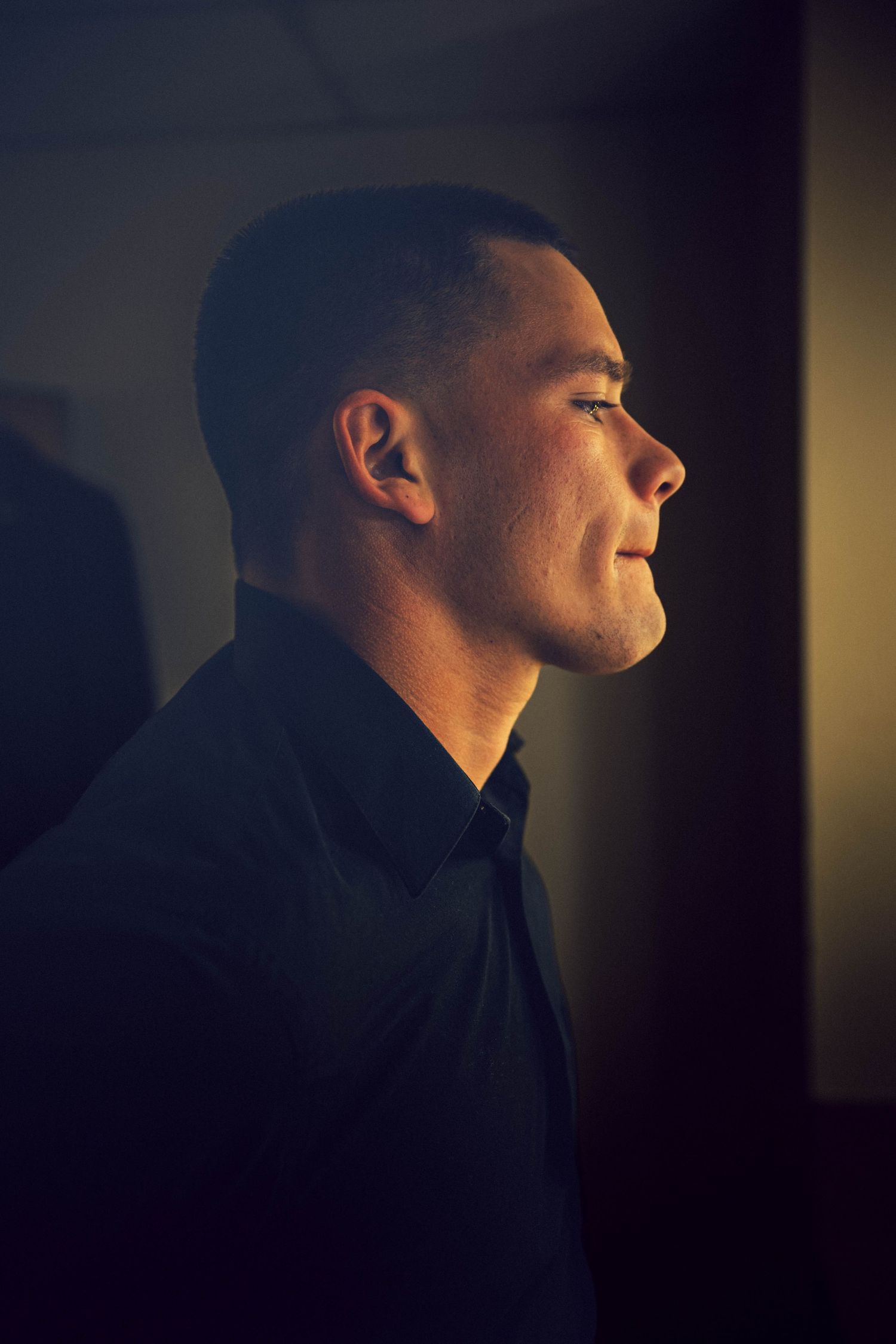 Side profile of a short-haired man in a dark button-up shirt, lit by soft warm light against a dim, moody background.