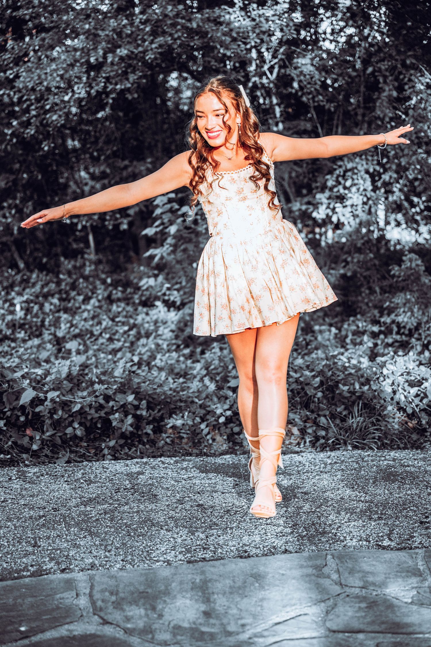 Smiling young woman with long wavy hair twirls in a floral sundress and lace-up sandals on a stone path, set against lush green trees.