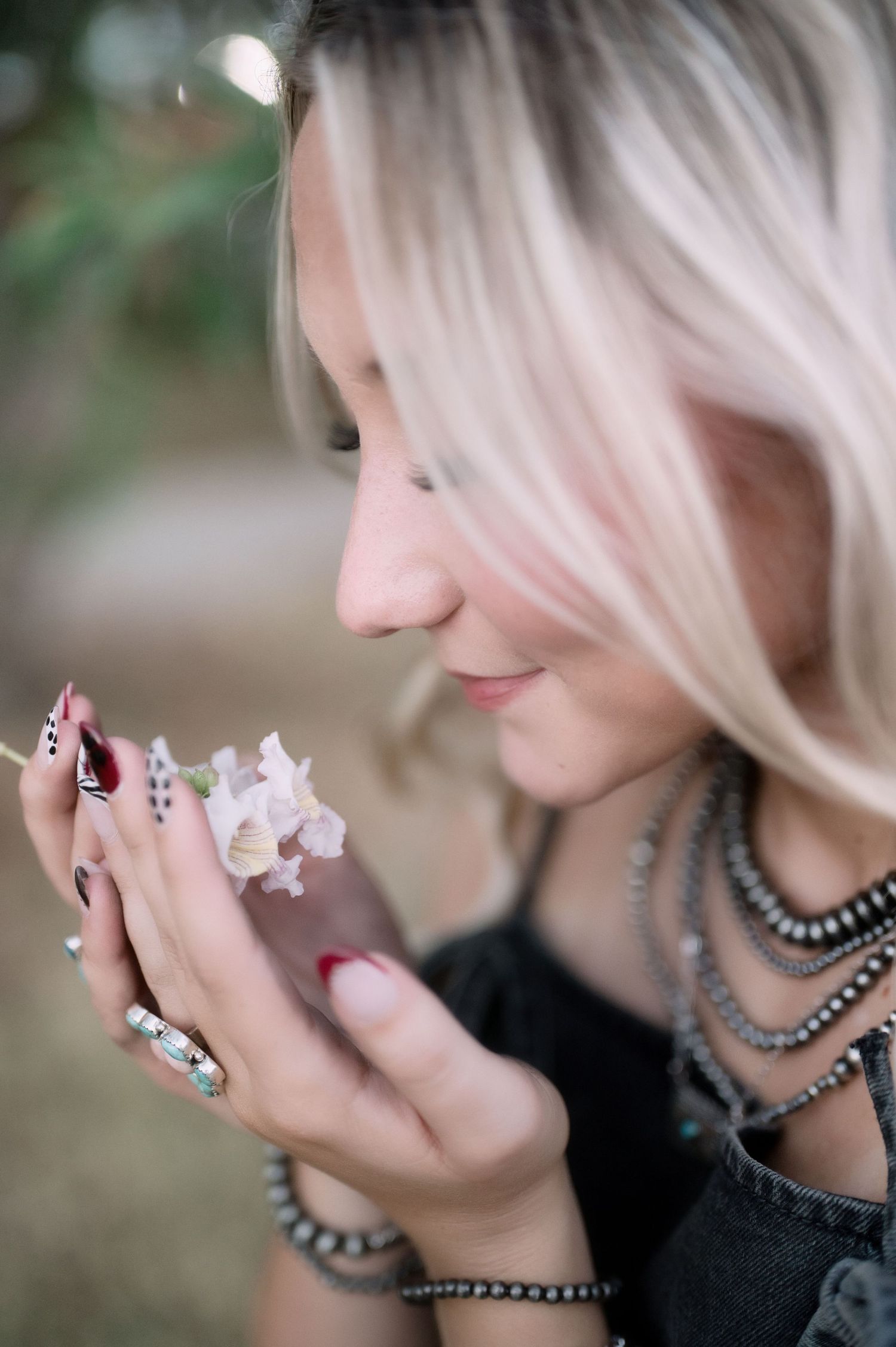 Blonde woman with red nails and layered beaded necklaces smelling a small pink flower outdoors.
