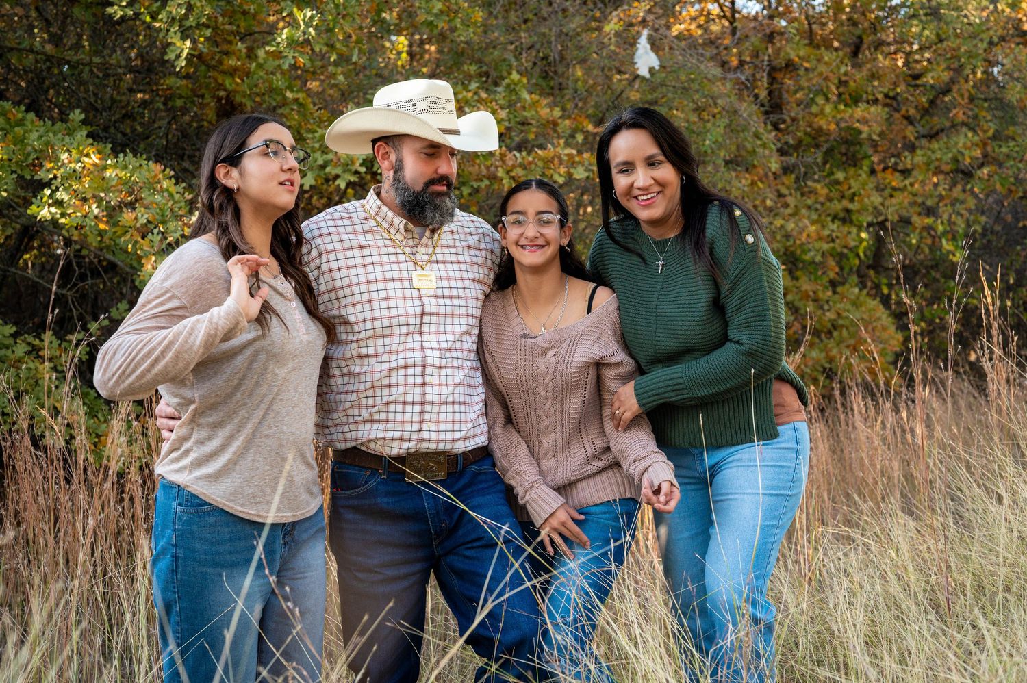 A family laughs together while posing outdoors in a field with autumn foliage in the background. Azle family photo session.