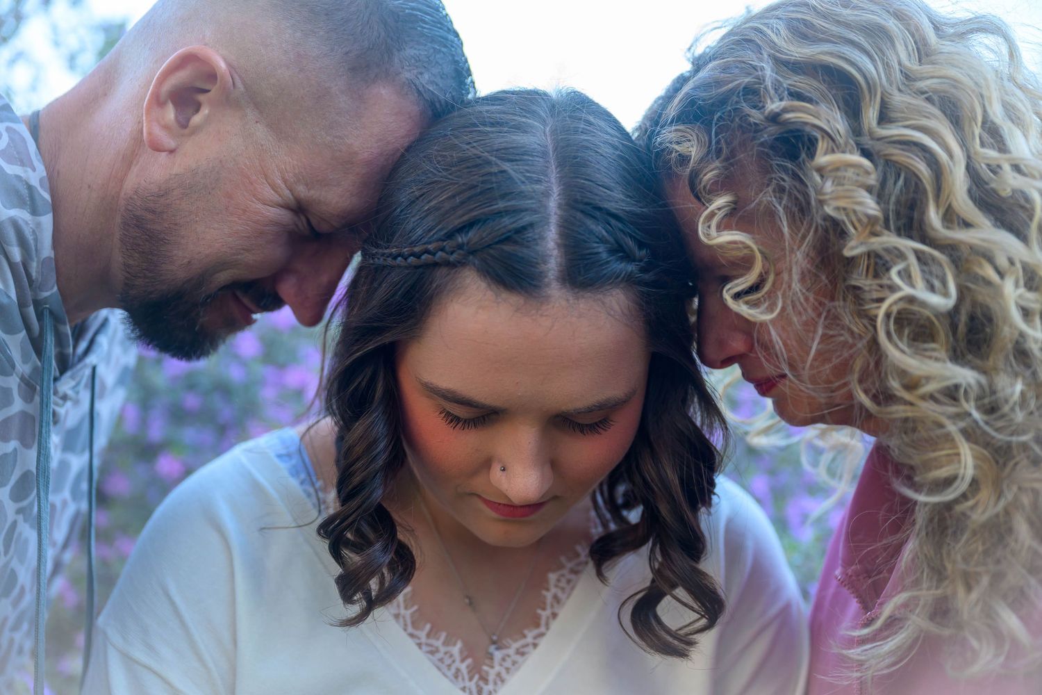 Three people stand close with foreheads touching and eyes closed in a quiet, emotional moment, with blurred purple flowers behind them.